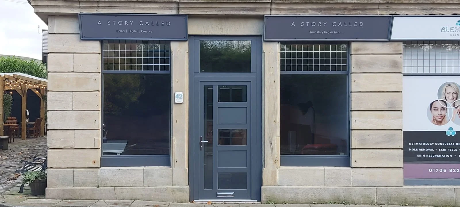 Exterior view of 'A Story Called' storefront with gray door and windows, adjacent Blemish Clinic signage, and outdoor seating area.