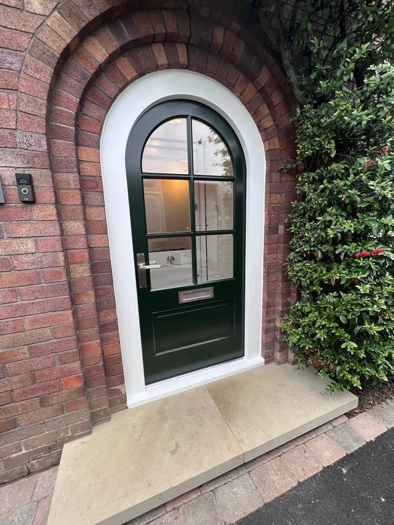 Arched black front door with glass panes, surrounded by red brick wall and greenery.