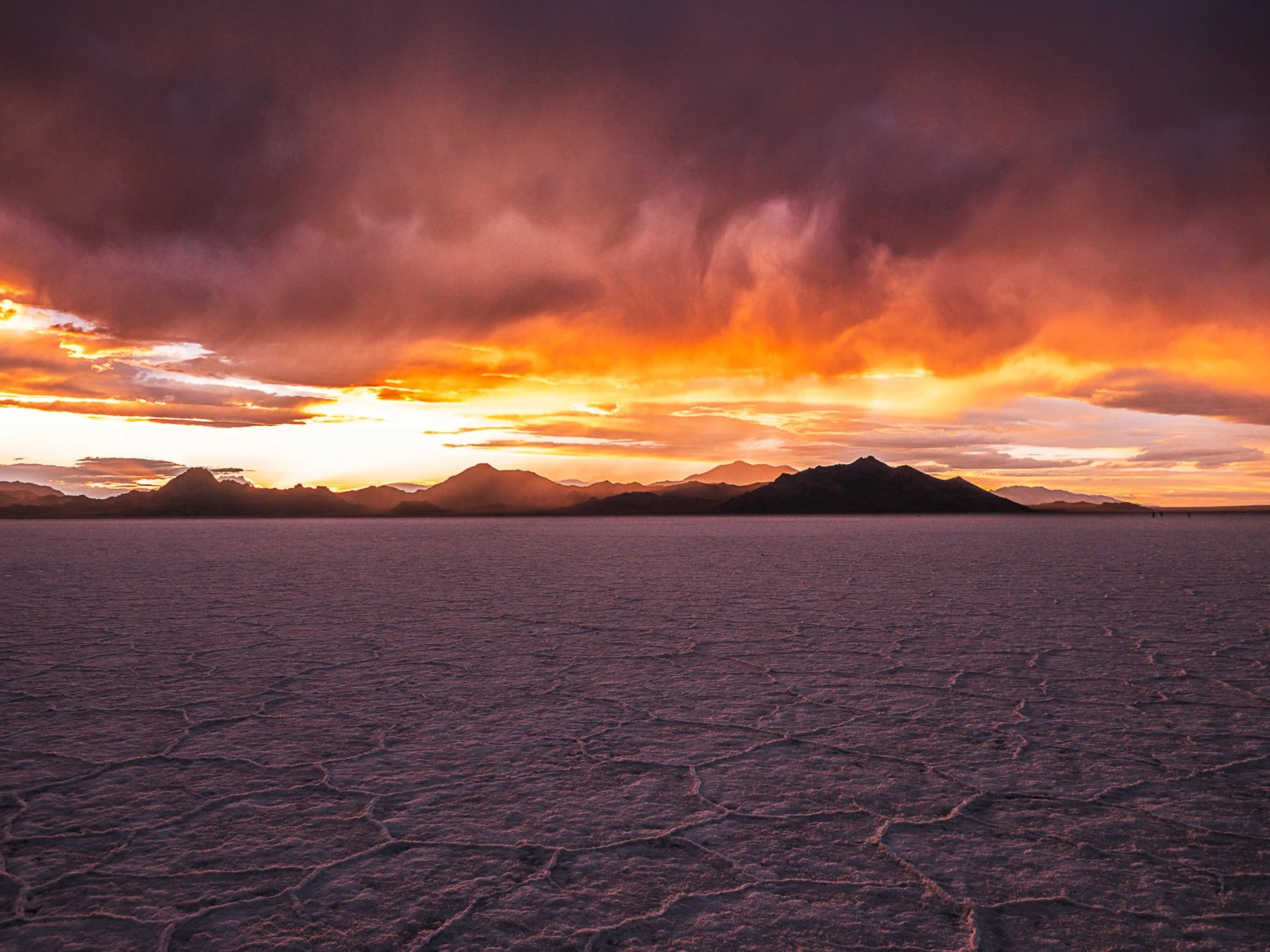 Bonneville Salt Flats are shown during a firey sunset behind the mountains with the colors of the sunset reflecting onto the storm clouds above and rock hard dry salt flats themselves.