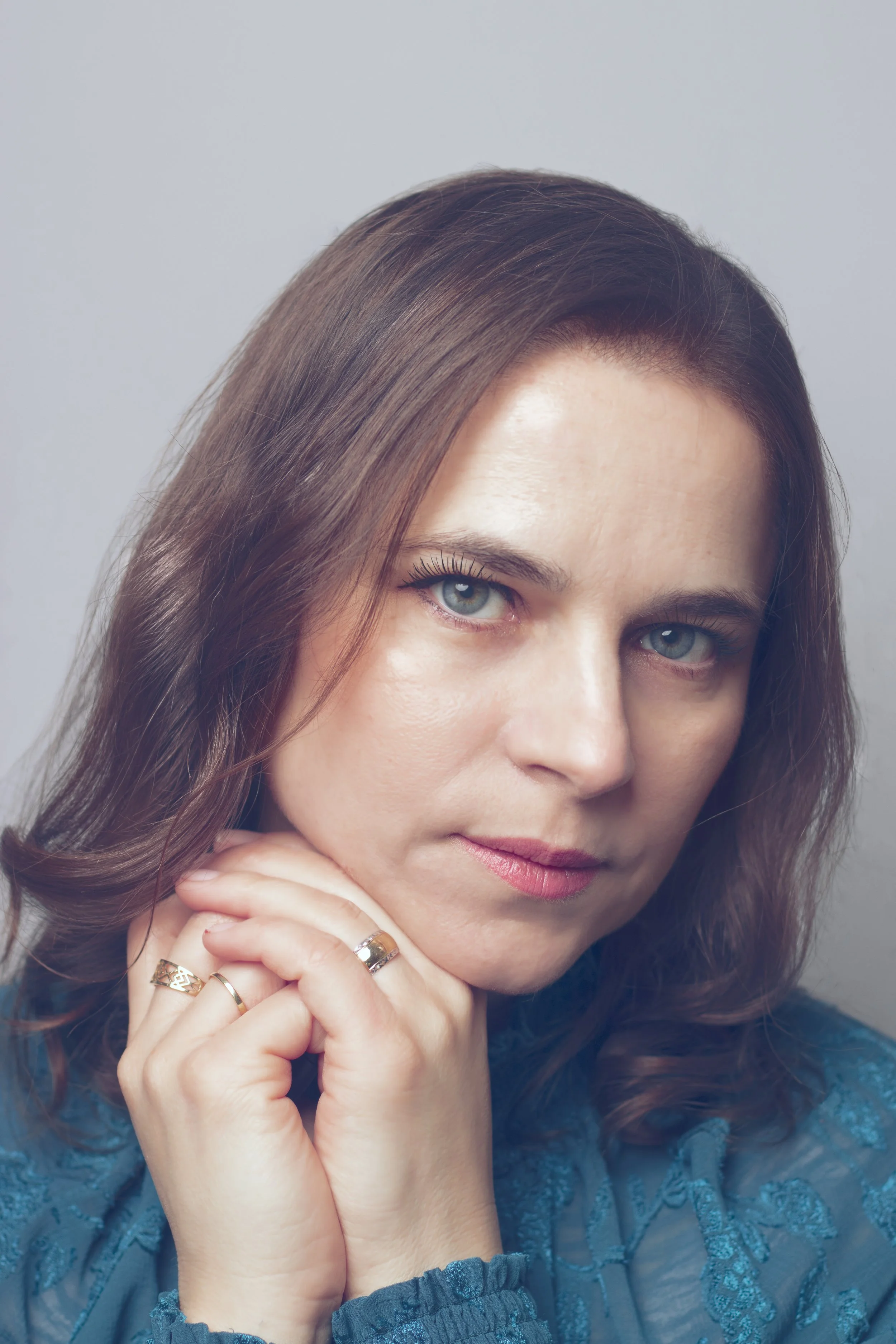 Close-up of a woman with brown wavy hair, blue eyes, light makeup, wearing rings, and a blue lacy dress.