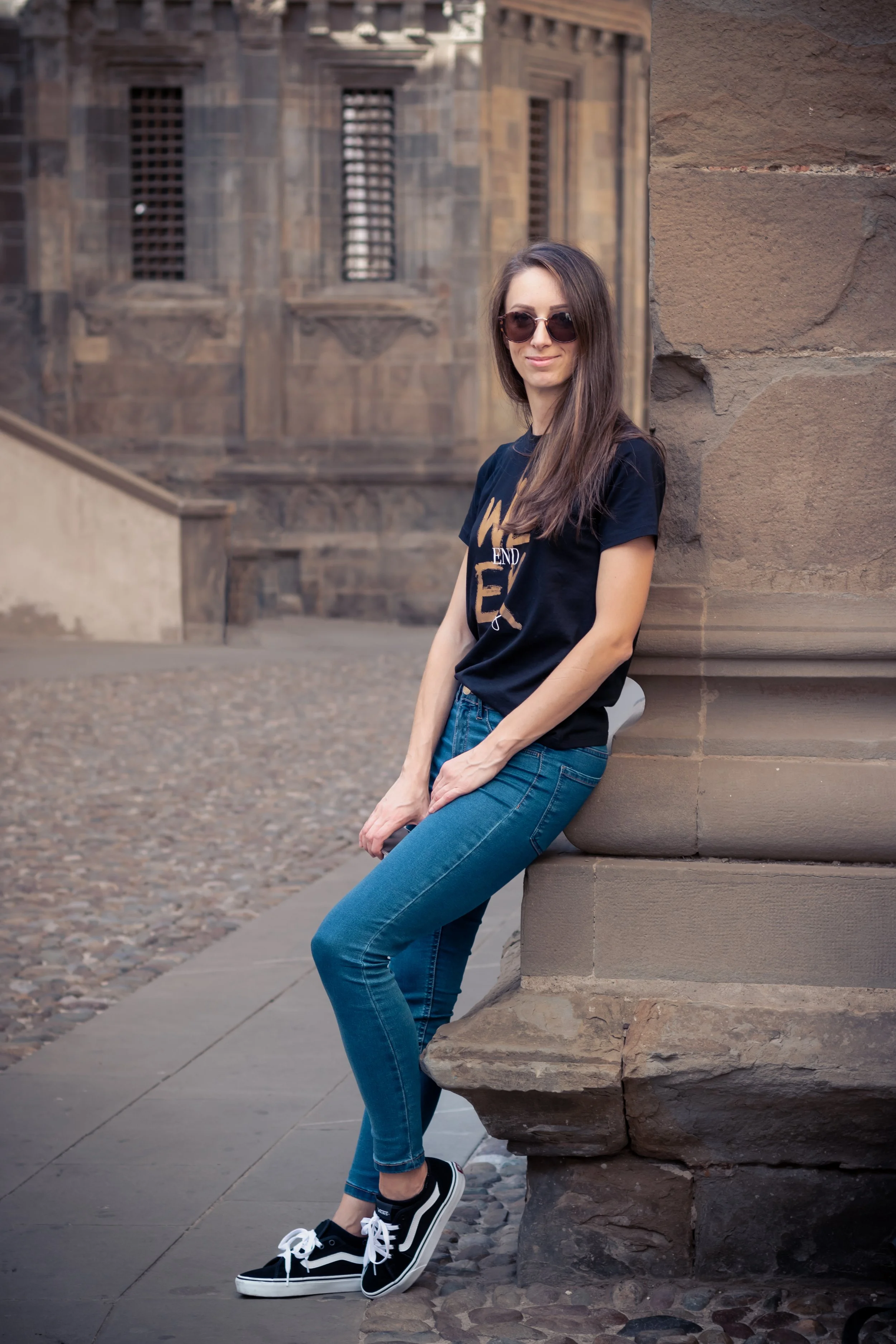 A woman wearing sunglasses, a black t-shirt, and blue jeans leaning against a brick wall on a city street.