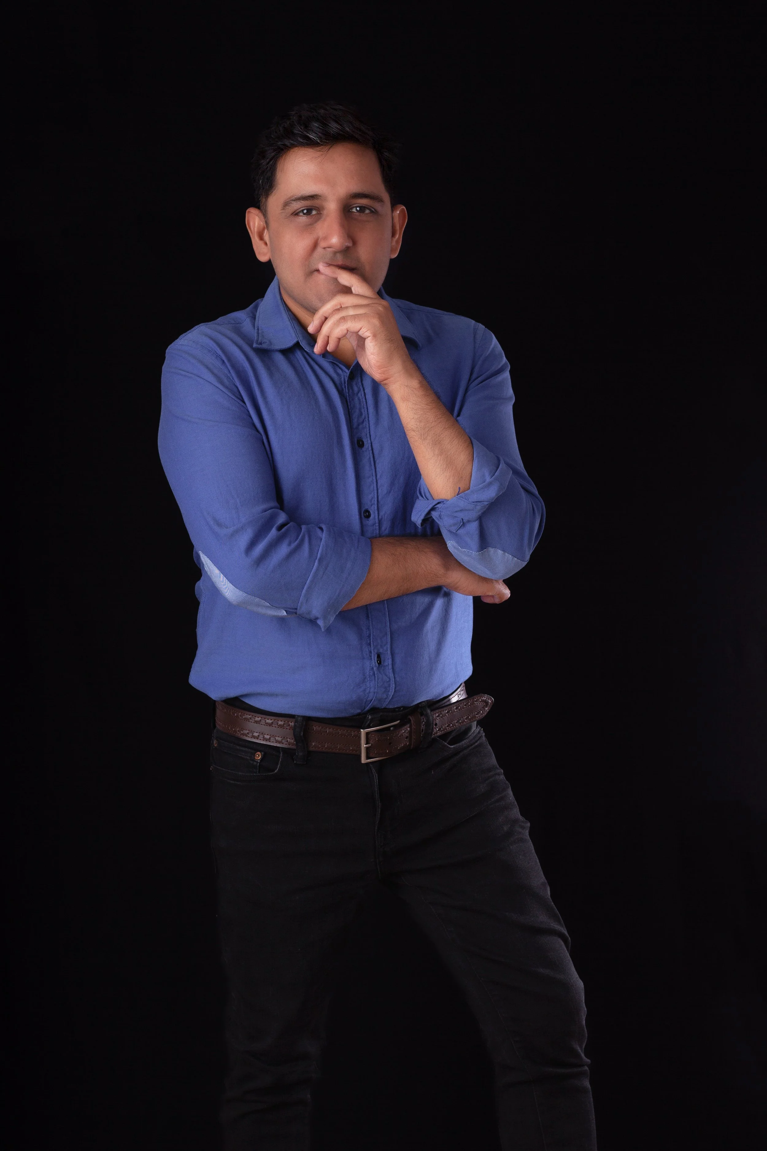 A young man wearing a blue shirt and black pants poses against a black background, with one arm crossed and the other hand touching his lips.