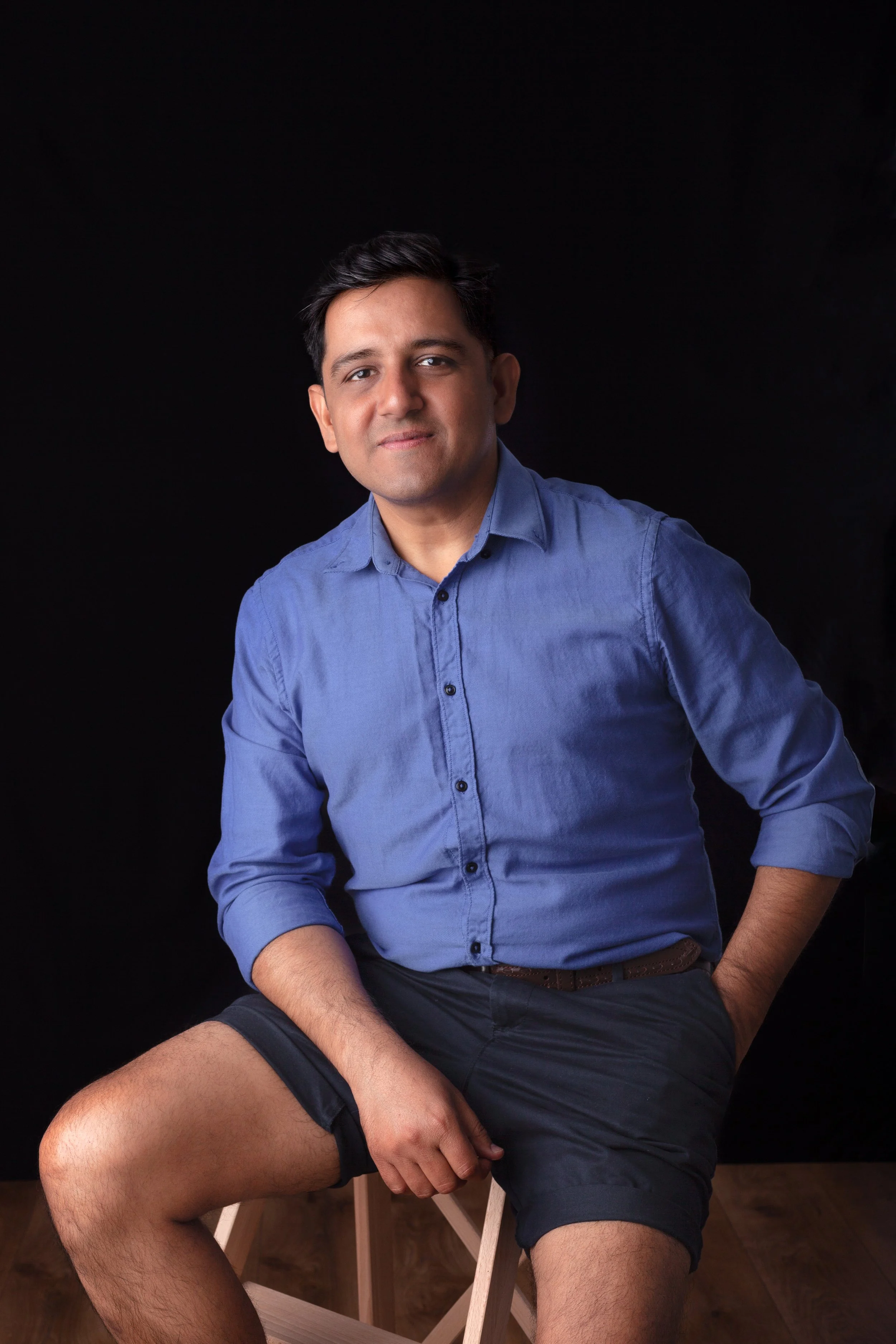 A young man with dark hair and light skin, wearing a blue button-up shirt and dark shorts, sitting on a wooden chair against a black background. He has a confident expression, with one arm resting on his knee and the other on his waist.