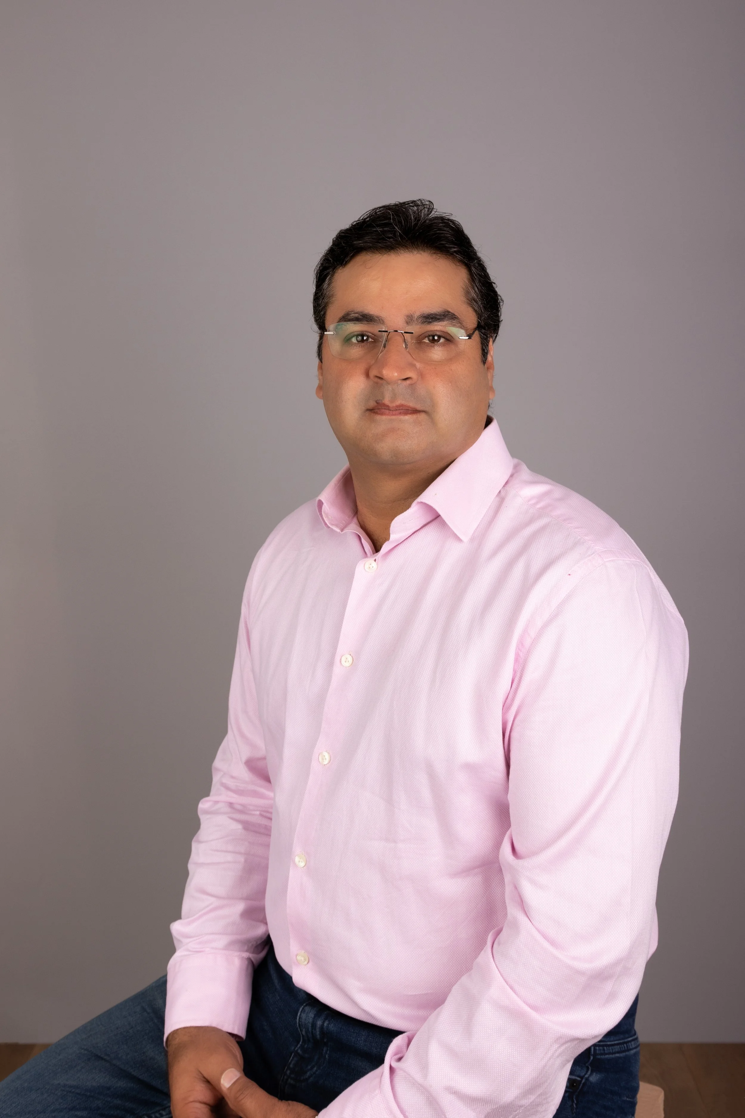 A man with dark hair and glasses wearing a pink button-up shirt, seated against a gray background.