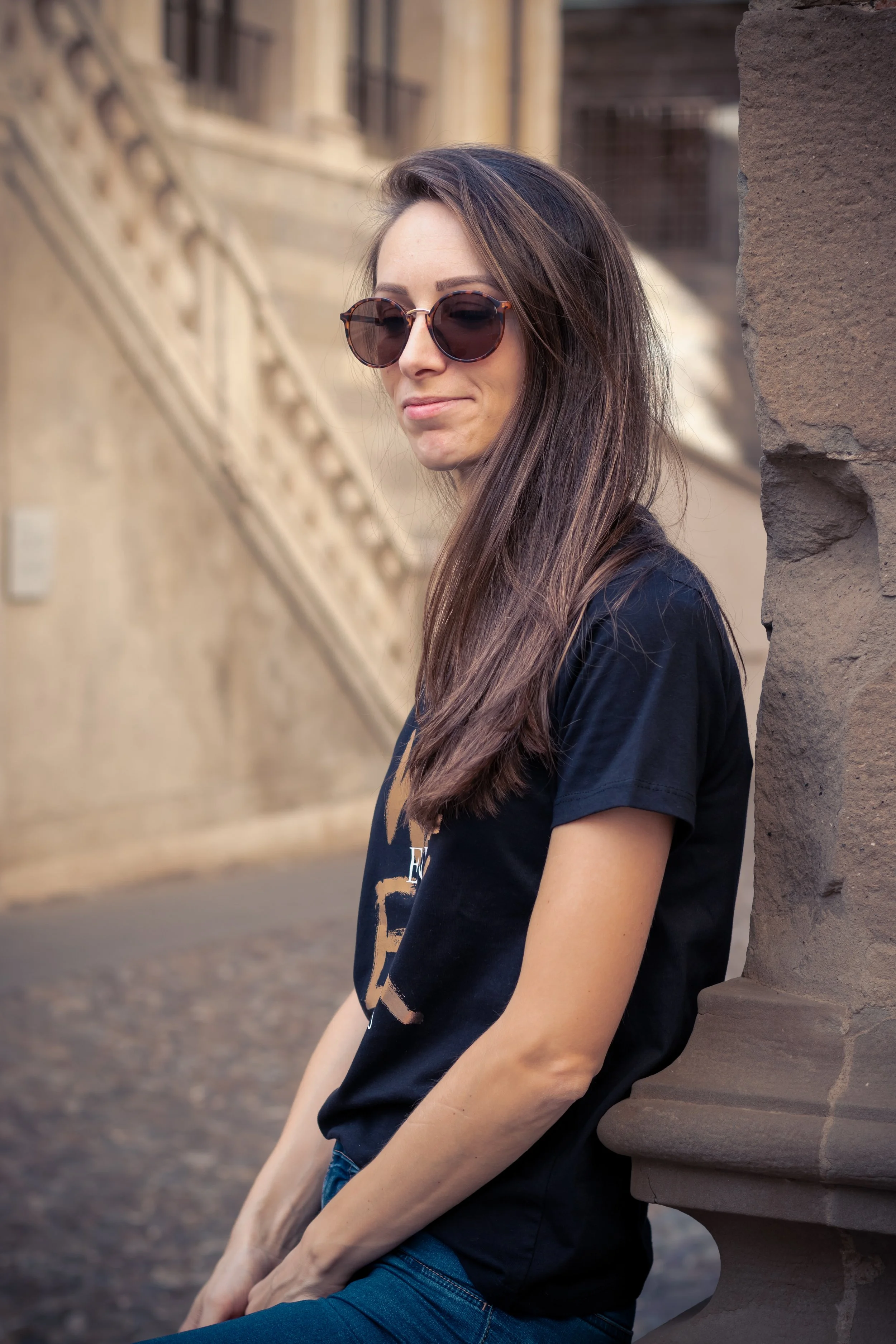 A woman with long brown hair wearing sunglasses and a black T-shirt with a beige graphic, sitting against a stone monument or wall outdoors during daytime.