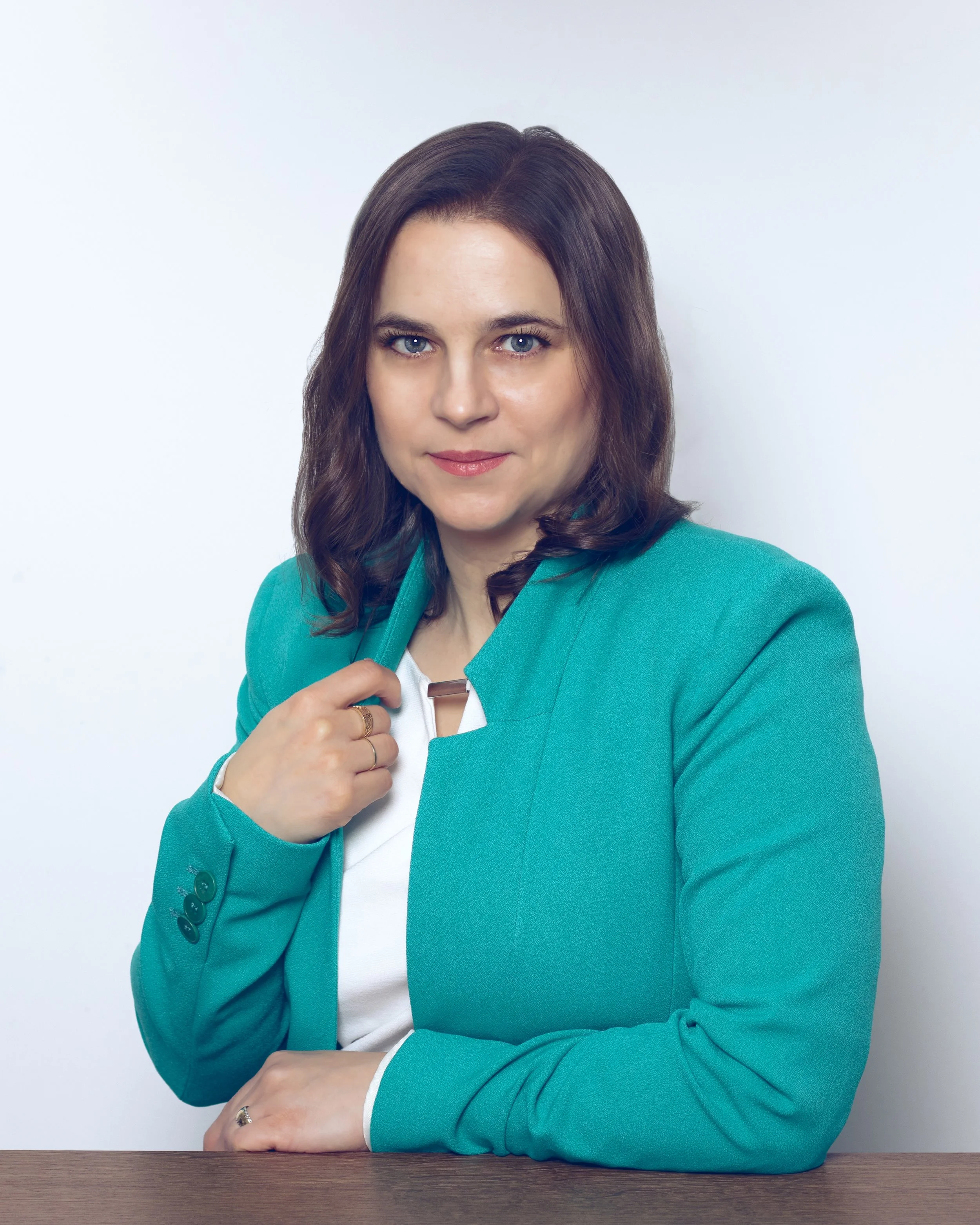 A woman with long brown hair and blue eyes wearing a teal blazer, white shirt, gold ring, and holding a metal clip, sitting at a wooden table against a plain white background.