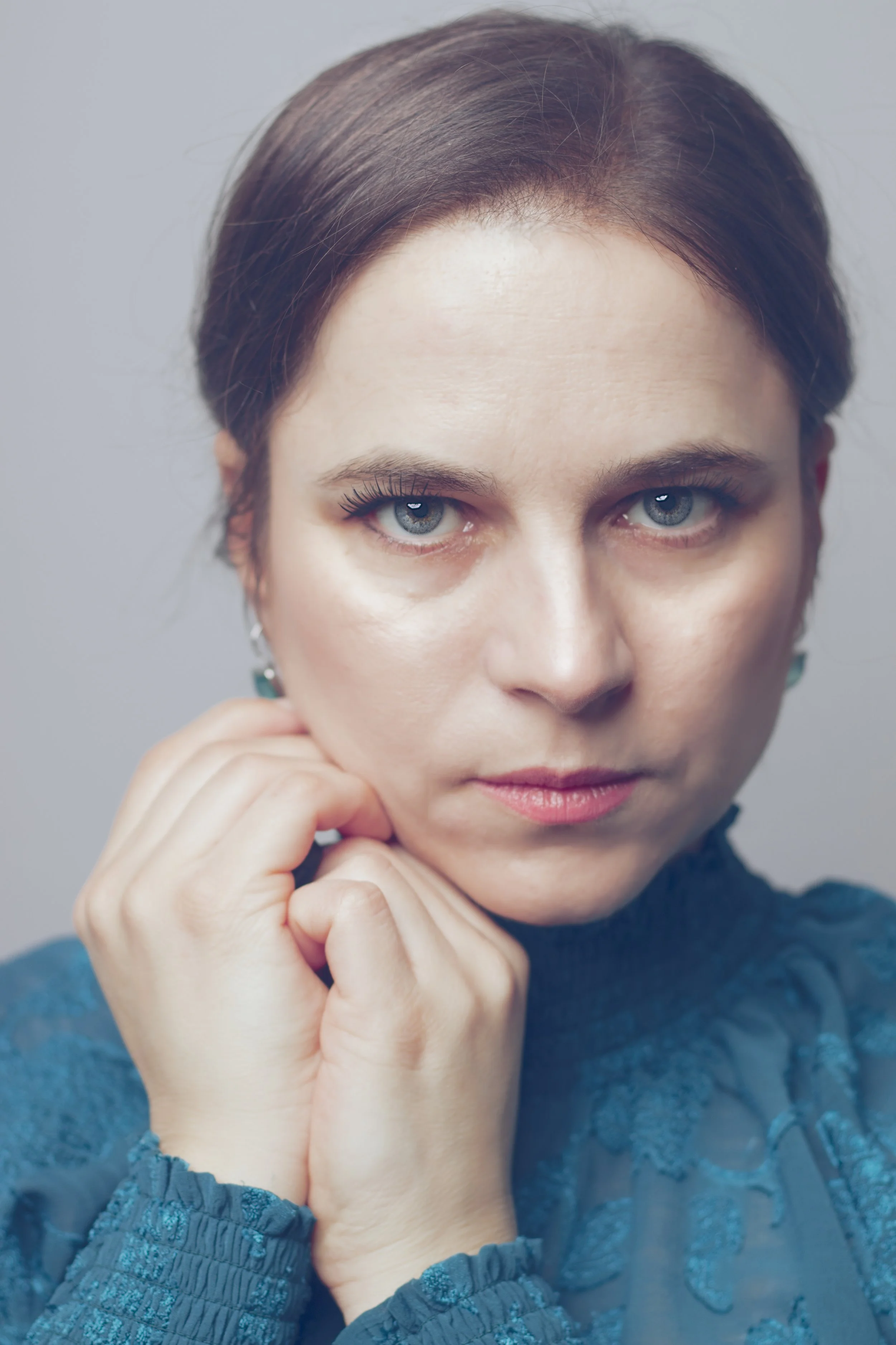 Close-up portrait of a woman with platinum blonde hair and blue eyes, wearing a blue textured sweater and earrings, with her hands resting near her face.