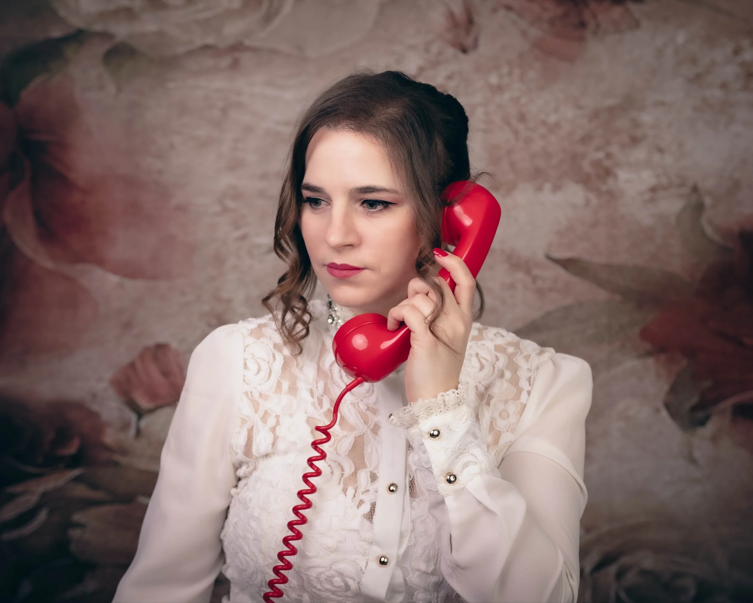 A woman with brown hair and makeup holding a red vintage telephone receiver to her ear, wearing a white lace dress with pearl embellishments, standing in front of a textured stone wall.