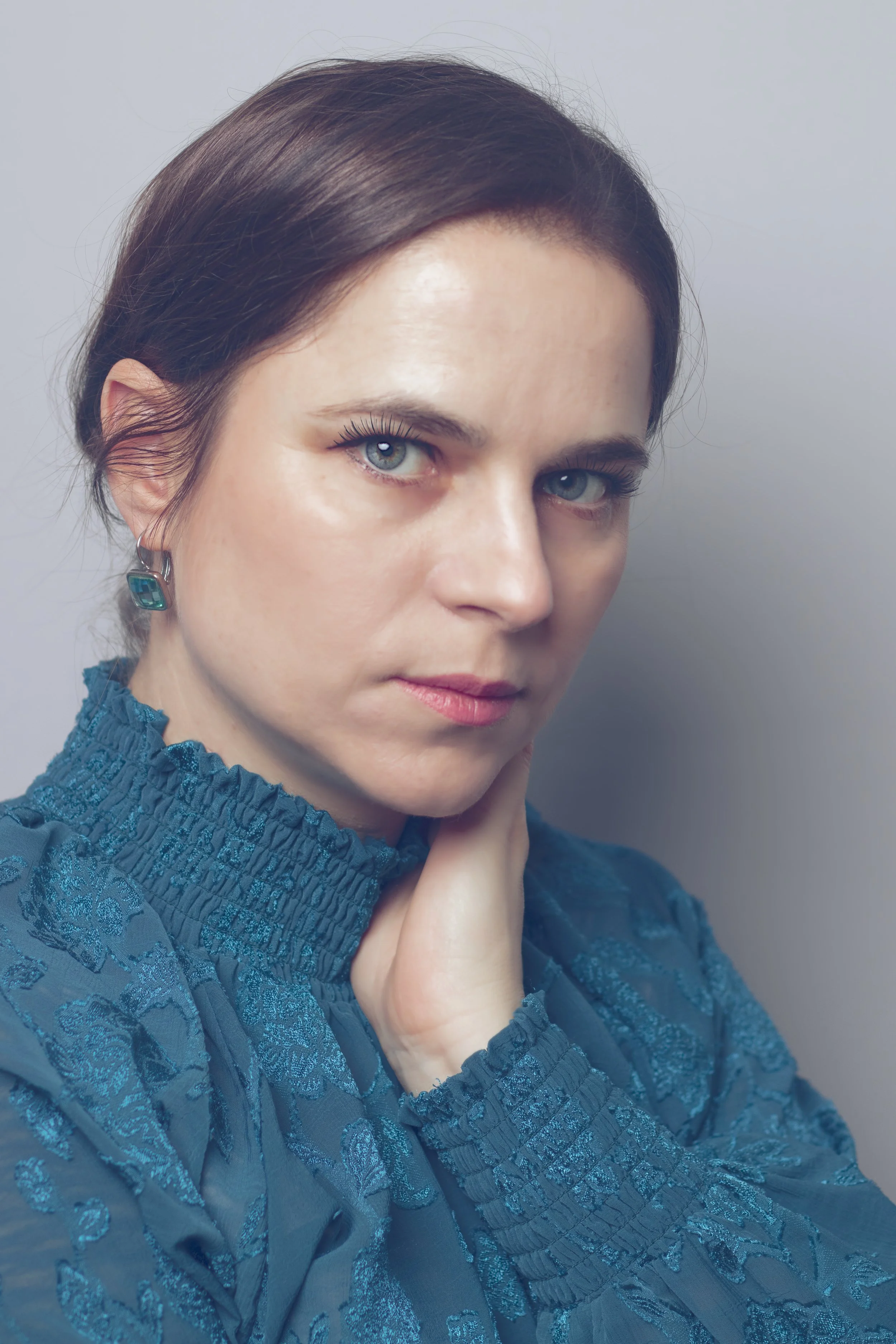 A woman with dark brown hair and blue eyes wearing a blue textured blouse and a turquoise earring, resting her face on her hand against a plain gray background.