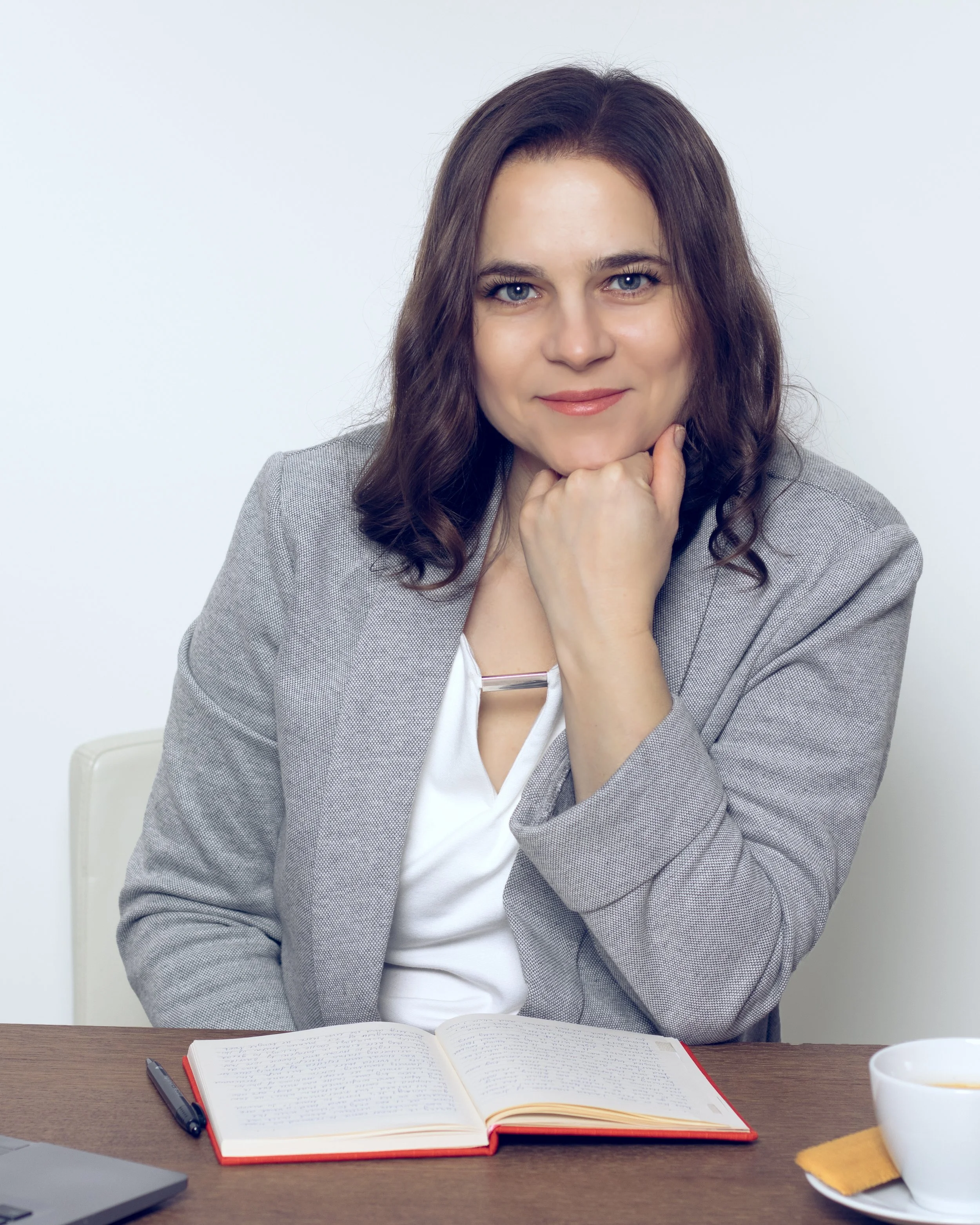 A woman with wavy brown hair and blue eyes sitting at a desk with an open notebook, a pen, a cup of coffee, and a yellow cloth. She is resting her chin on her hand and looking at the camera.