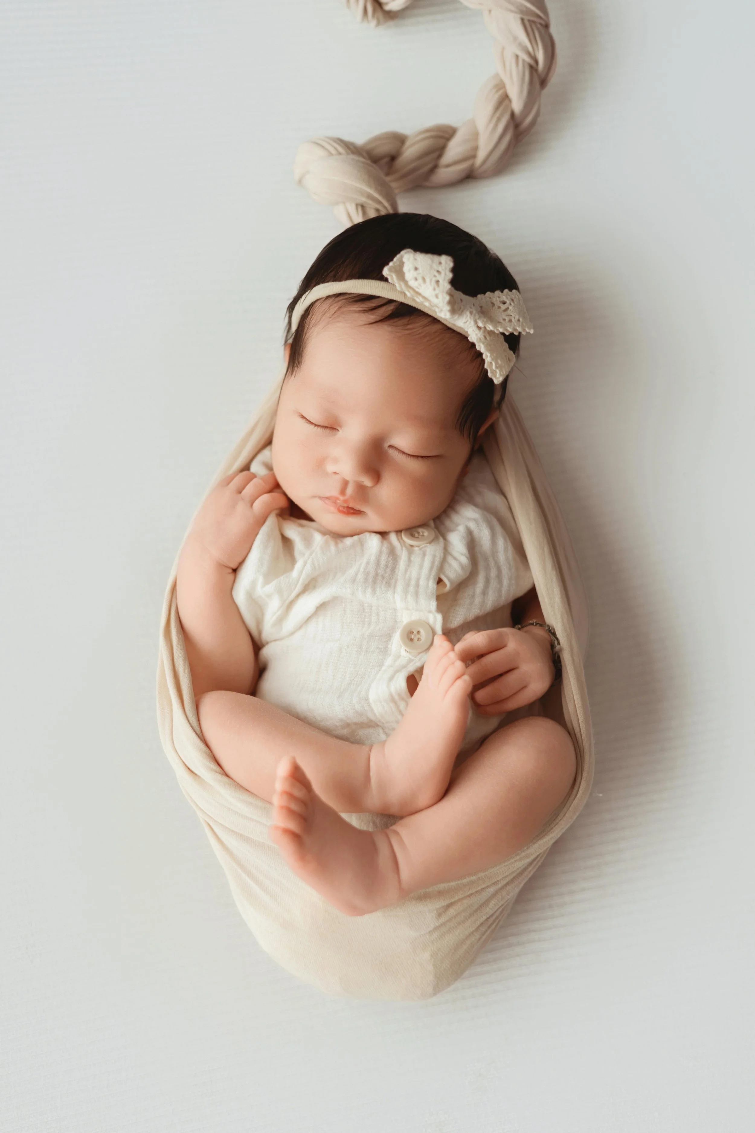 A sleeping baby with dark hair, wearing a white headband with a lace bow, wrapped in a beige blanket, lying on a white surface.