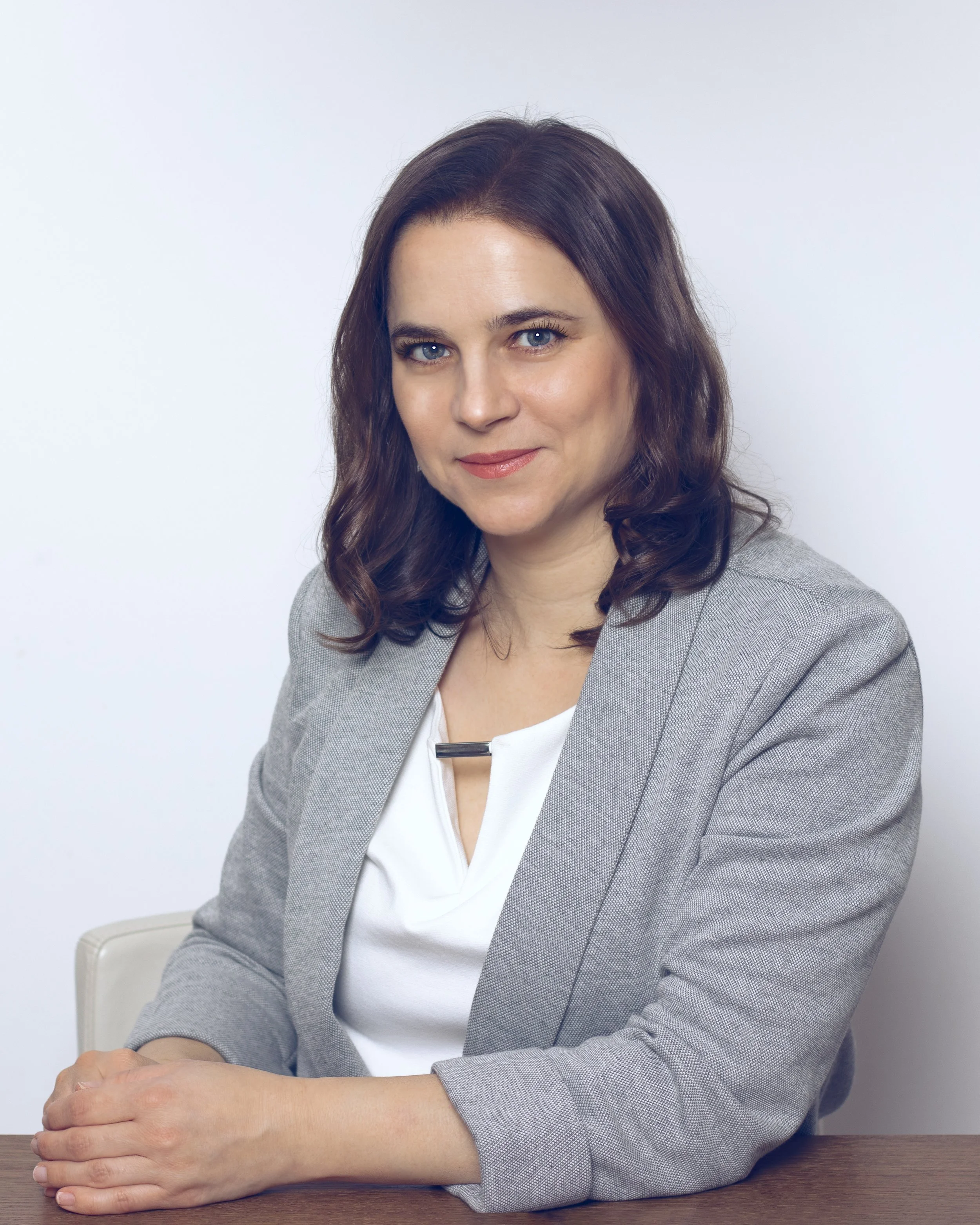 A woman with shoulder-length brown wavy hair and blue eyes, wearing a light gray blazer and a white top, sitting at a wooden table with her arms crossed. The background is plain white.