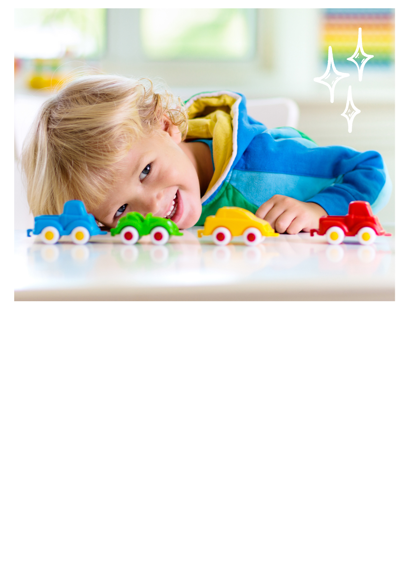 Smiling toddler in a blue hoodie resting their head on a table beside a row of colourful toy cars