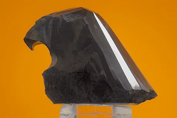 The Black Falcon, a 612.34-carat black carbonado diamond shaped like a falcon's head, displayed against an orange background. Conceptual rendering by The Jeweler Blog.