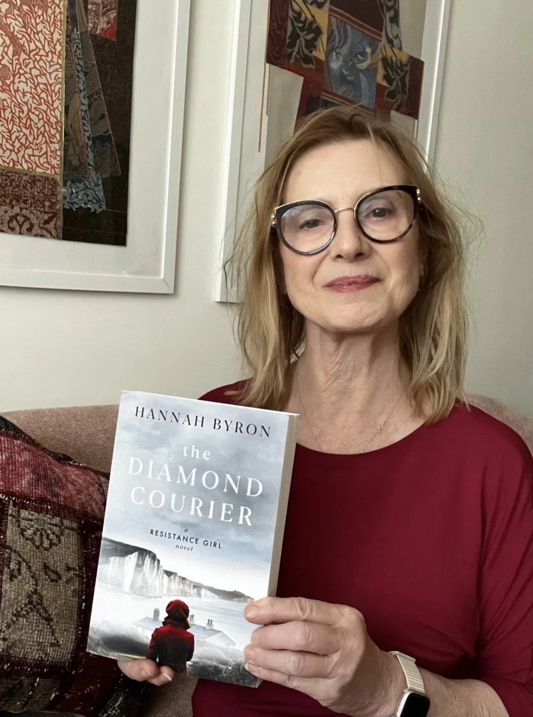 Author Hannah Byron smiling and holding a paperback copy of her novel The Diamond Courier, a Resistance Girl novel, in front of framed artwork.