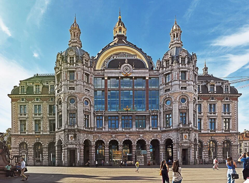 The ornate stone façade of Antwerp Central Station on a sunny day, with its grand dome, turrets and arched glass window rising above the station square.