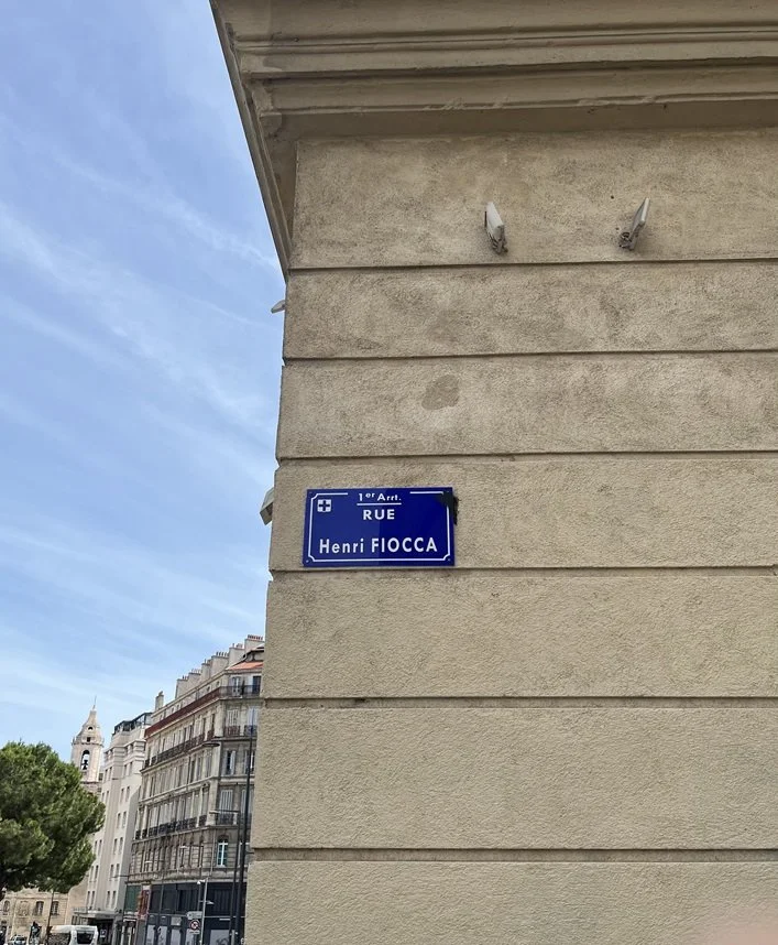 Street sign showing Rue Henri Fiocca on the side wall of a building with several classical styled buildings in the background and a clear blue sky above them