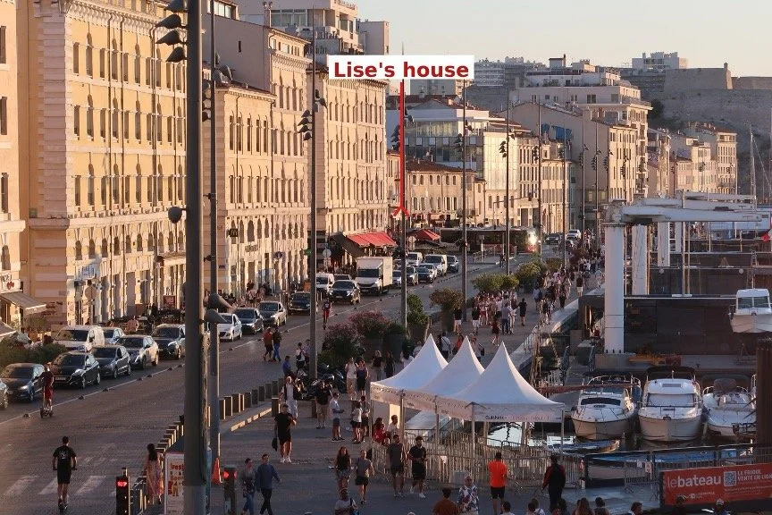The Quai de Rive Neuve on Marseille's Vieux Port in the evening sun, busy with people, cars and boats. A red arrow marks number 15, the house where SOE agent Lise de Baissac lived for decades after the war.