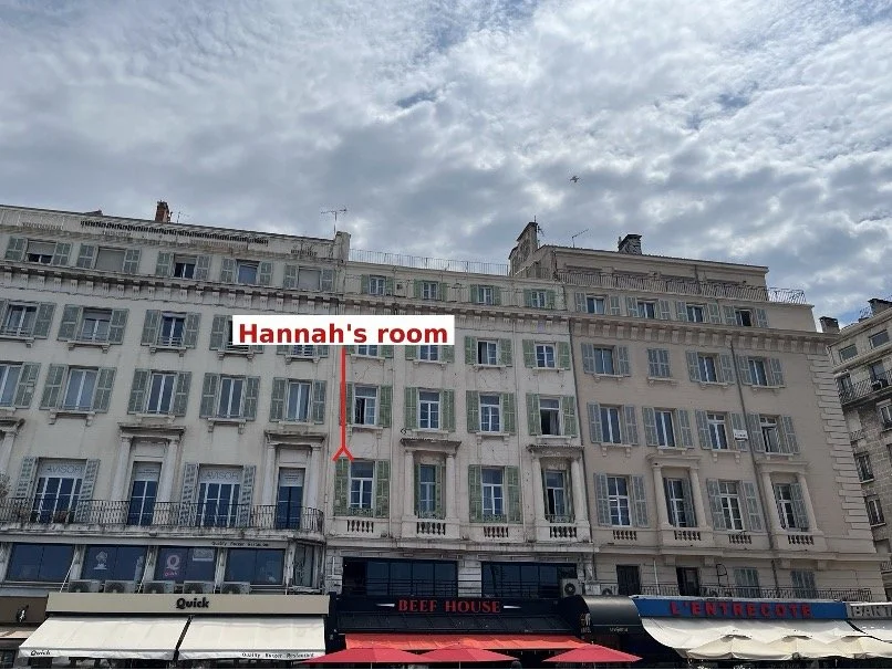 A hotel on Marseille's Vieux Port with pale shutters and restaurant awnings below. A red arrow marks the window of author Hannah Byron's room, from which she could see Lise de Baissac's house across the harbour.