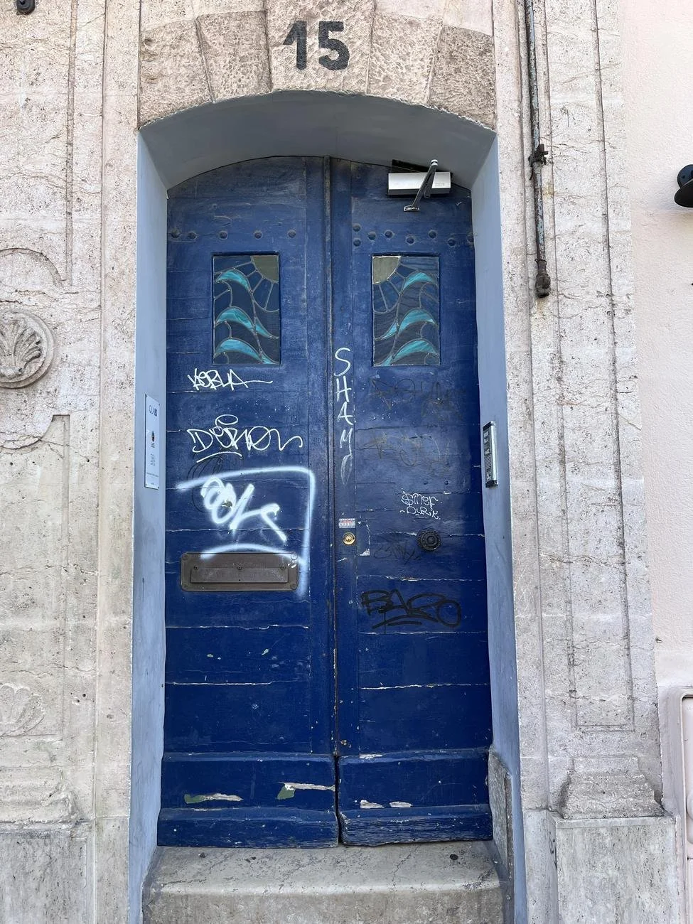 The old blue double front door of 15 Quai de Rive Neuve, with stained-glass panels and the number 15 on the stone arch above, covered in graffiti tags.