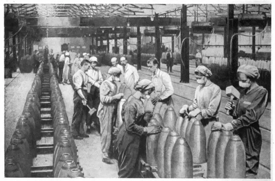 A black and white photograph of a British munitions factory, where women in overalls and headscarves, some wearing cloth masks, work along a row of large artillery shells alongside male workers.