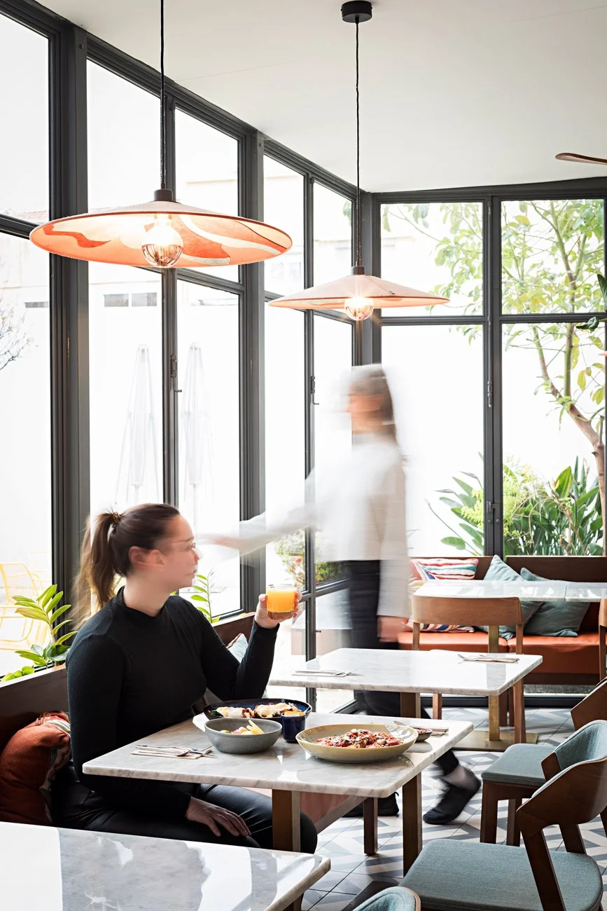 Cafeteria moderna com luz natural, mesa com pratos de comida e bebida, pessoa sentada segurando um copo, decoração com plantas e lâmpadas pendentes.