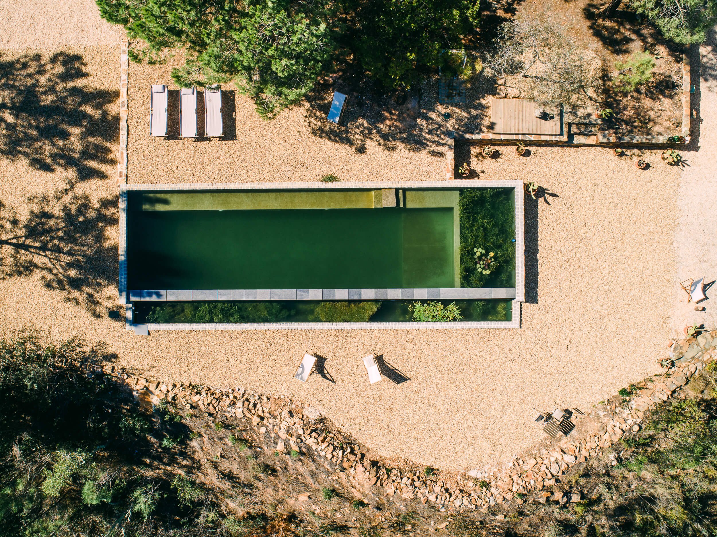 Vista aérea de uma piscina retangular rodeada por um jardim com árvores e arbustos. À esquerda, há espreguiçadeiras alinhadas e um caminho de pedras contornando a área. A iluminação natural cria sombras das árvores no piso de pedras.