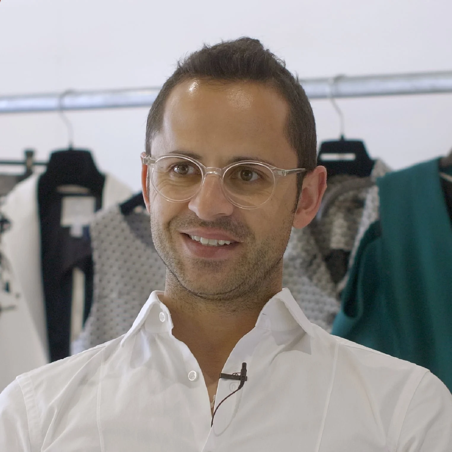 Homem usando óculos e camisa branca, com roupas ao fundo, em uma loja ou estúdio de moda.