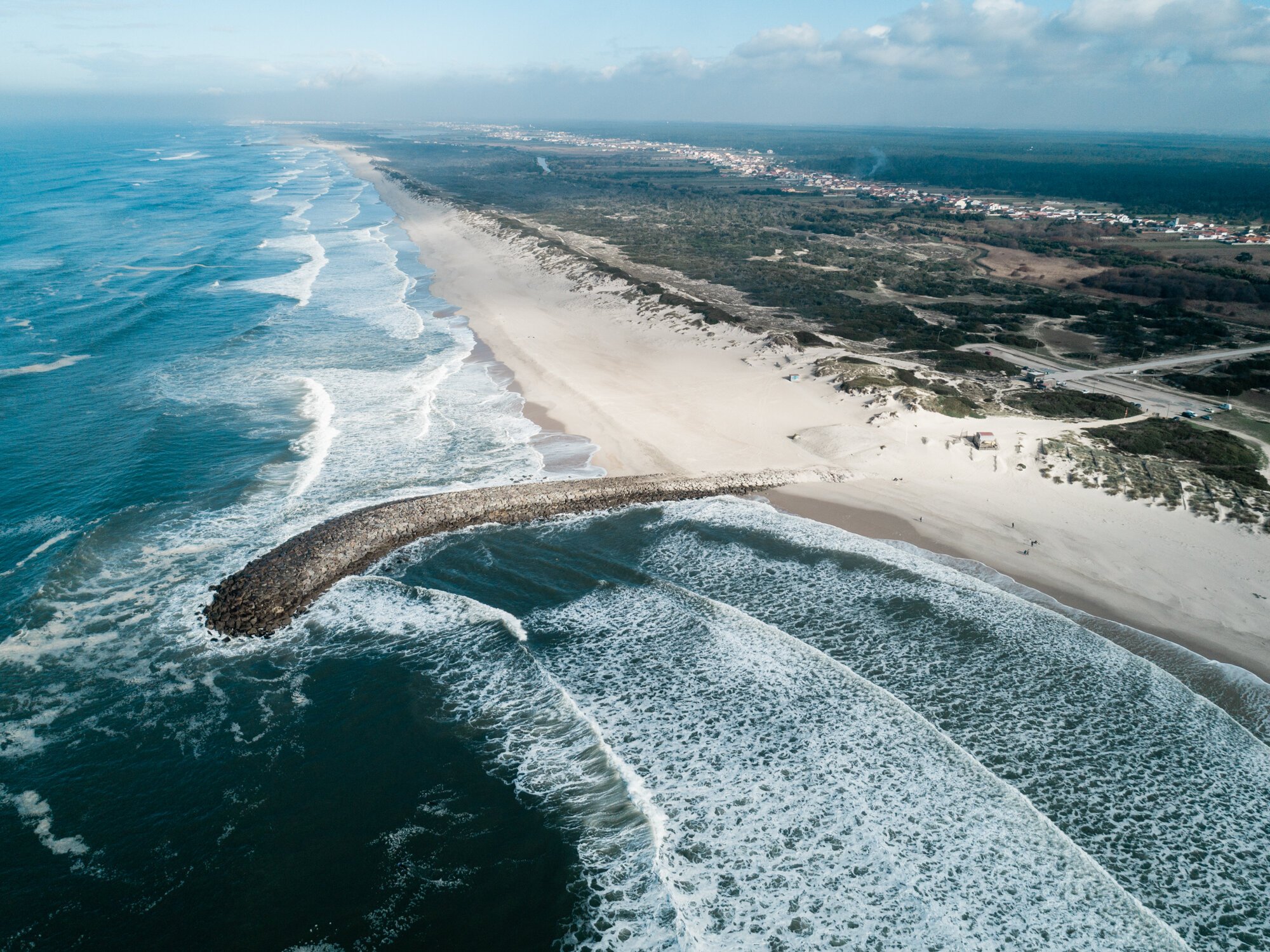 Vista aérea de uma praia com ondas quebrando e uma estrutura de rochas projetando-se no mar. A praia possui areia clara e densa vegetação nas proximidades. Céu parcialmente nublado.