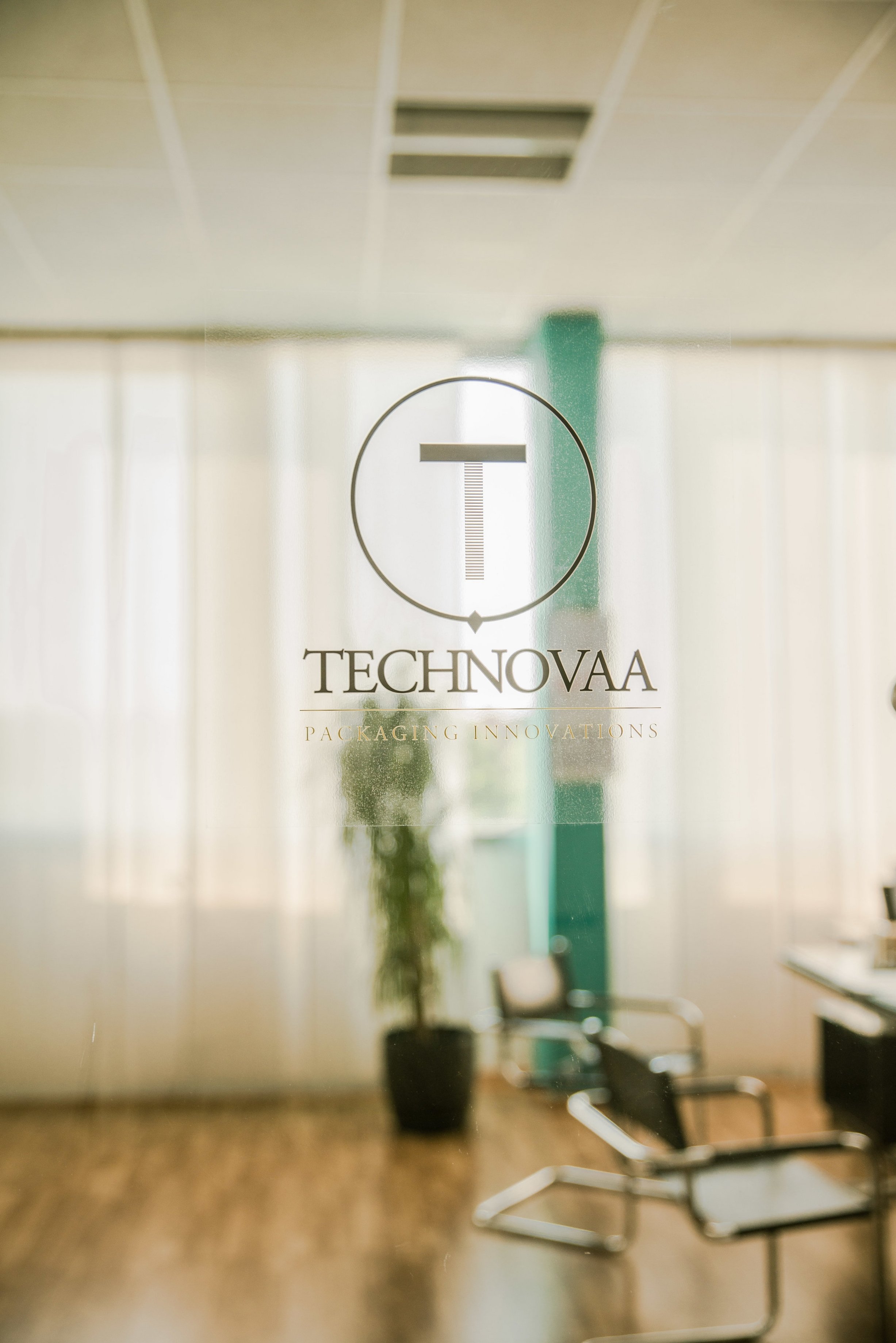 Logotipo "Technovaa Packaging Innovations" em vidro de uma porta de escritório, com sala de reuniões ao fundo.