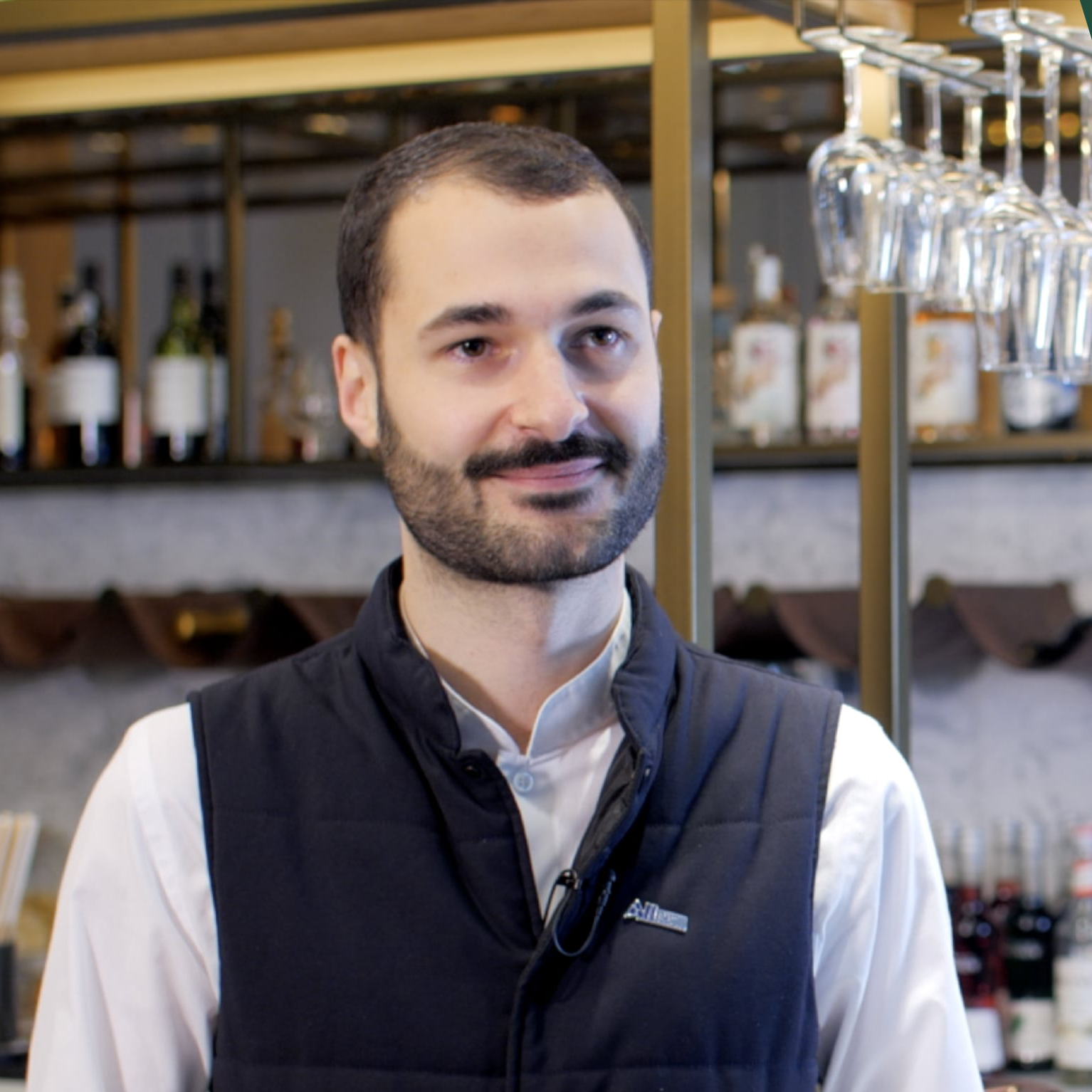 Homem sorrindo em um bar com garrafas de bebidas e taças penduradas ao fundo.