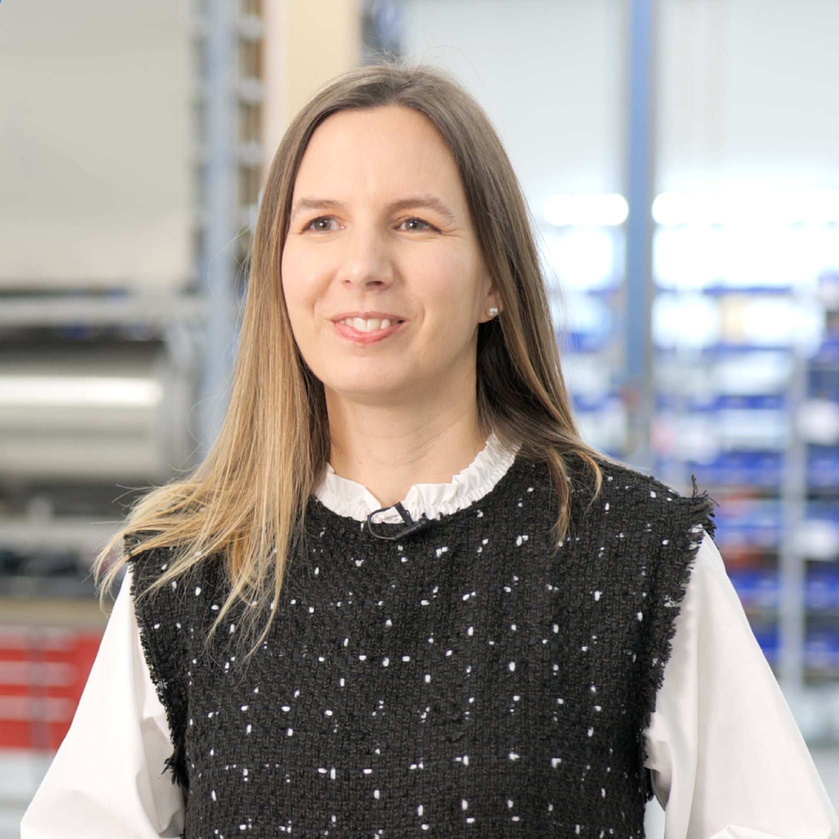 Mulher sorridente em um ambiente industrial, vestindo blusa preta com detalhes brancos e camisa branca.