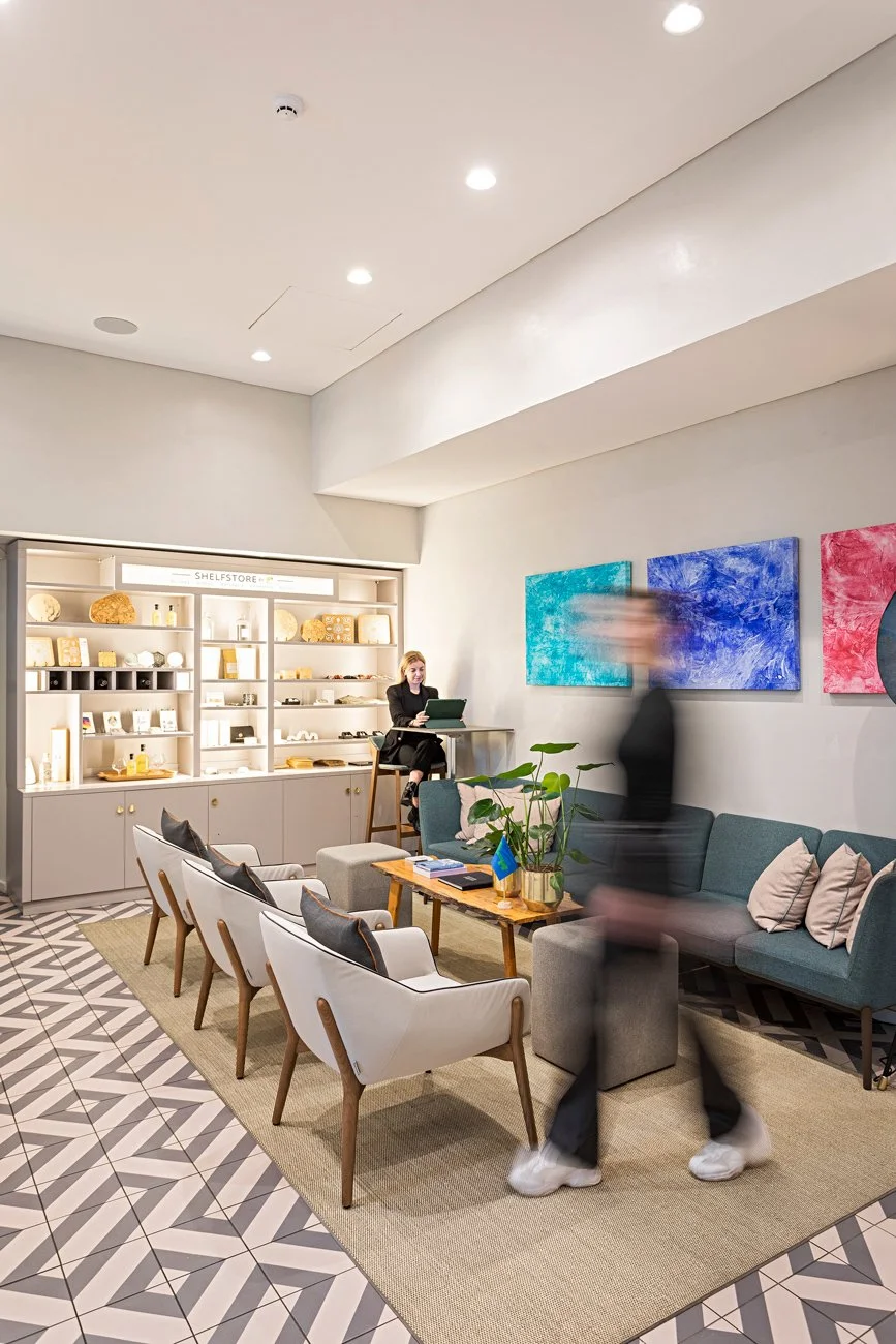 Sala de espera moderna com sofás, poltronas, mesa de centro, quadros coloridos na parede e estante iluminada com objetos. Pessoa andando em movimento borrado.