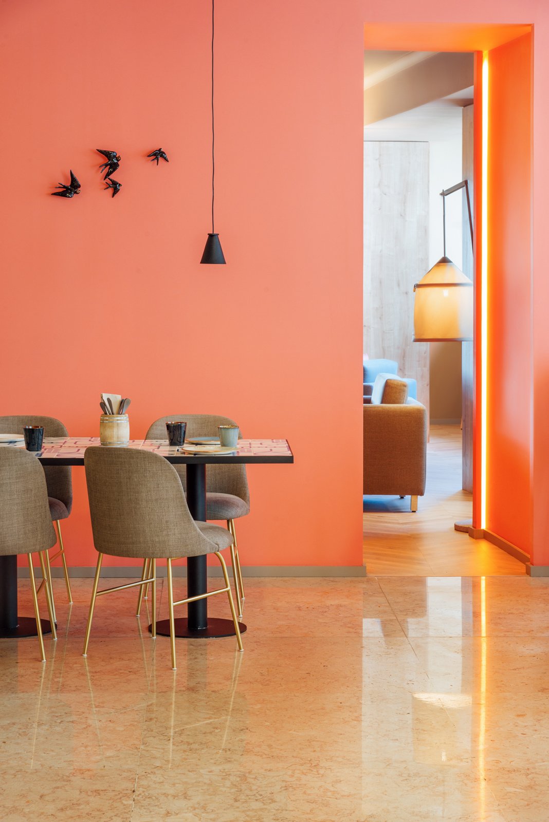 Sala de jantar moderna com parede laranja, mesa de jantar com cadeiras estofadas, luminária pendente preta, decoração de pássaros na parede e abertura para outra sala com sofás e luminária.