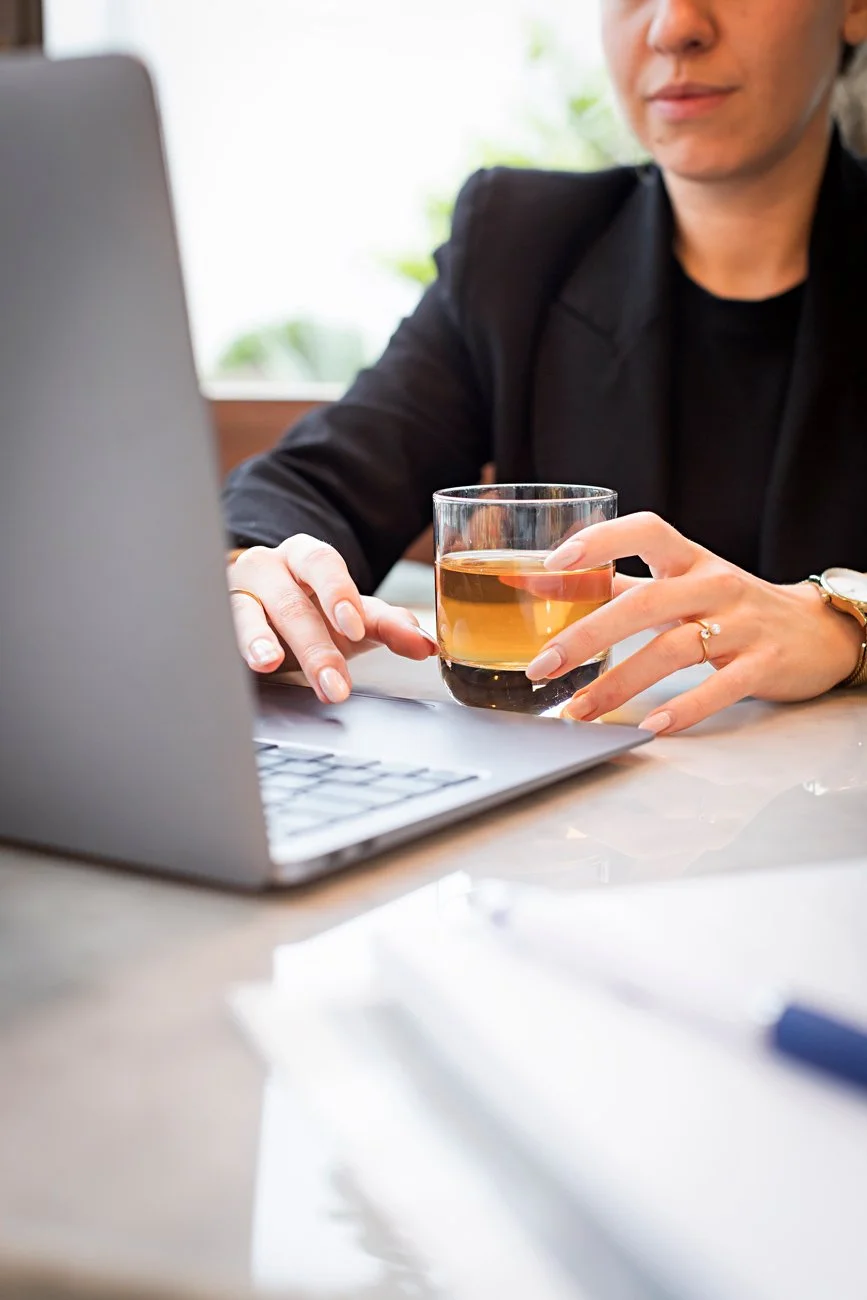 Pessoa com um copo de chá na mão em frente a um laptop.