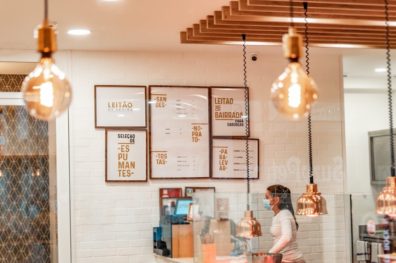 Restaurante com decoração moderna, incluindo lâmpadas pendentes. Mulher com máscara atrás do balcão. Placas na parede exibem o menu de "Leitão de Levira", destacando opções como "SANDES", "TOSTAS", e "LEITÃO À BAIRRADA". Parede de tijolos brancos ao