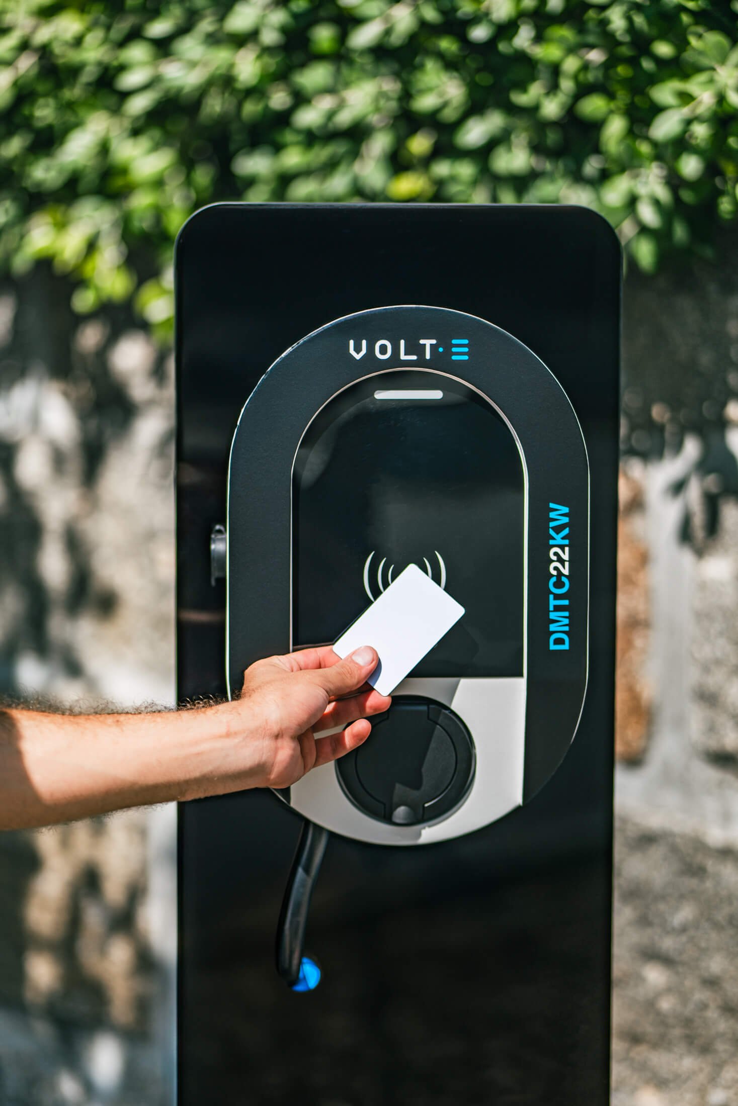 Estação de carregamento de carro elétrico com uma mão segurando um cartão RFID em frente ao leitor, em local externo com fundo de plantas.