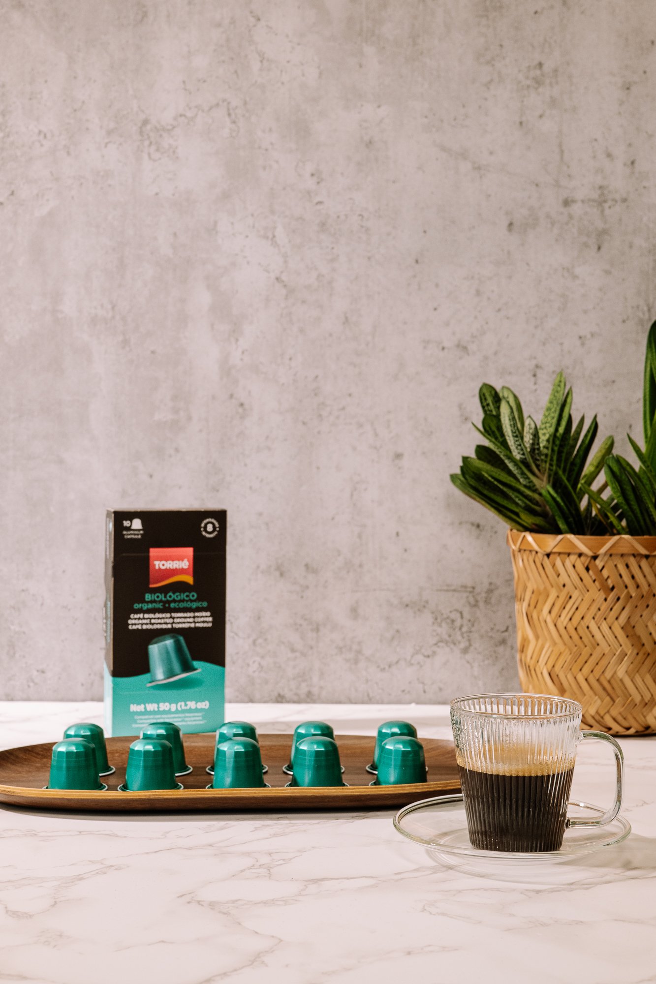 Cápsulas de café verde em um prato de madeira ao lado de uma embalagem com rótulo "Biológico" e uma xícara de café preto em um ambiente com uma parede de concreto e planta decorativa.