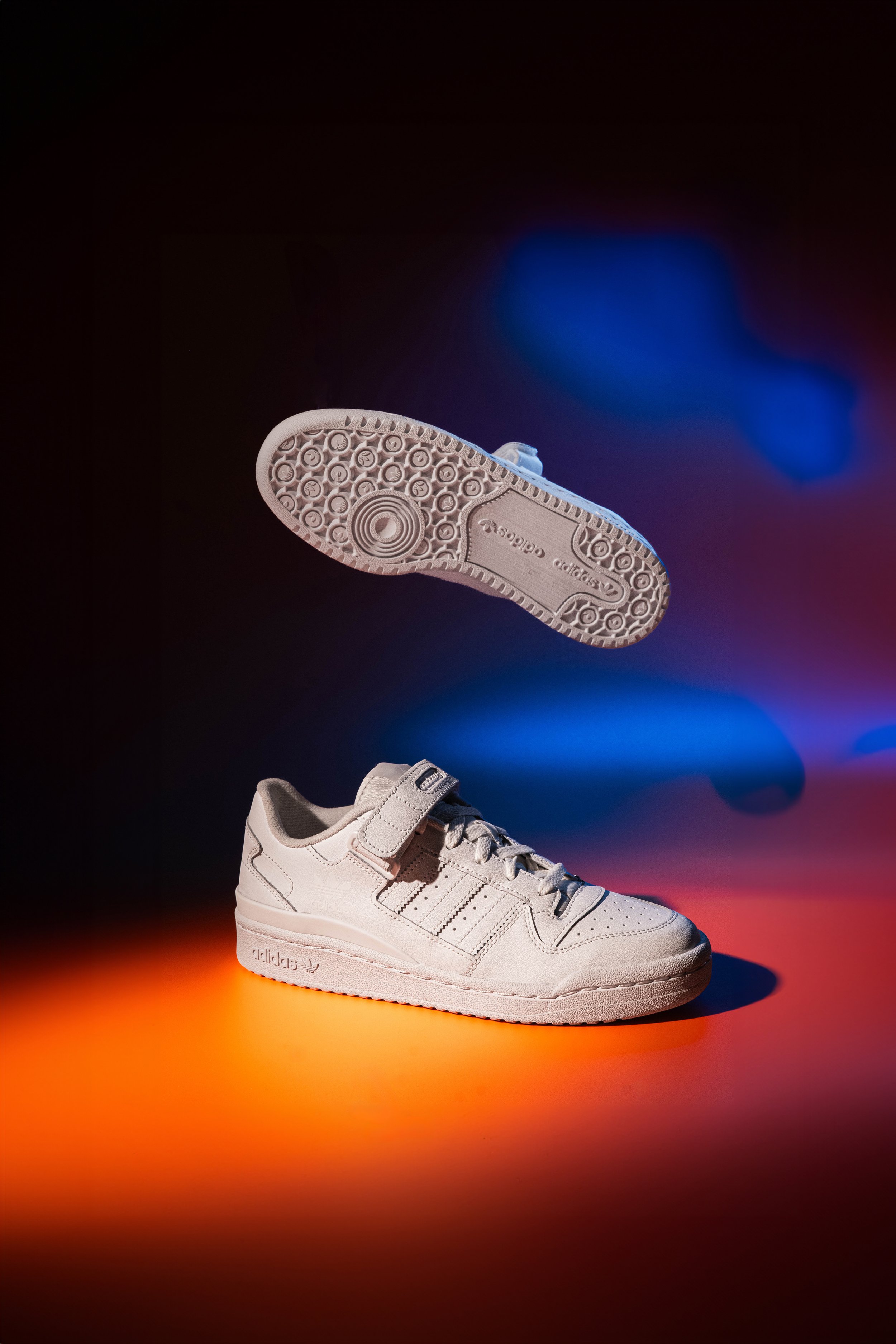 Tênis branco da marca Adidas em superfície colorida, com um em foco no solo e outro exibindo a sola no ar.