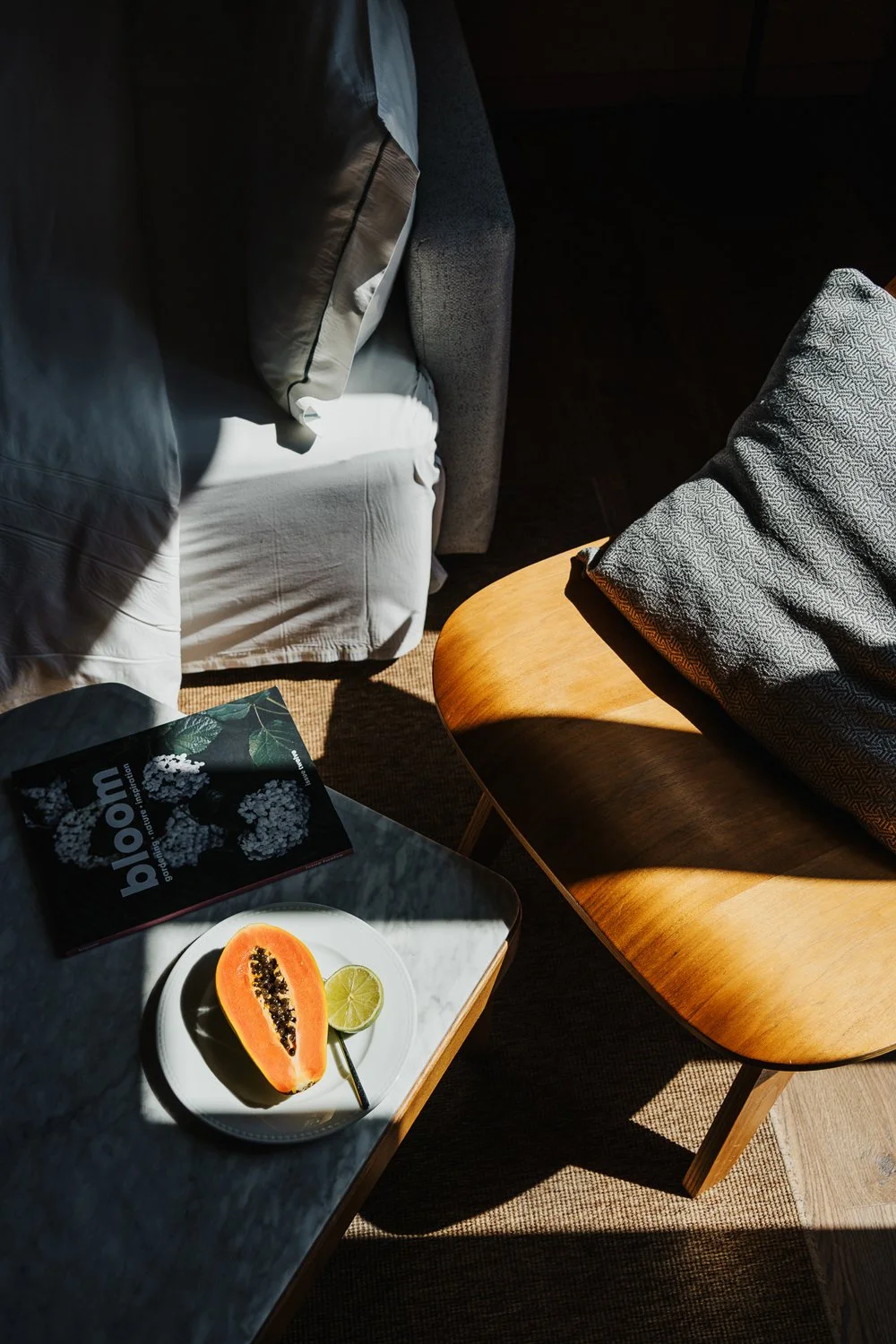 Metade de uma papaya e uma fatia de limão em um prato branco sobre uma mesa de mármore, com uma revista ao lado e uma poltrona com almofada ao fundo. A cena está iluminada por luz natural.