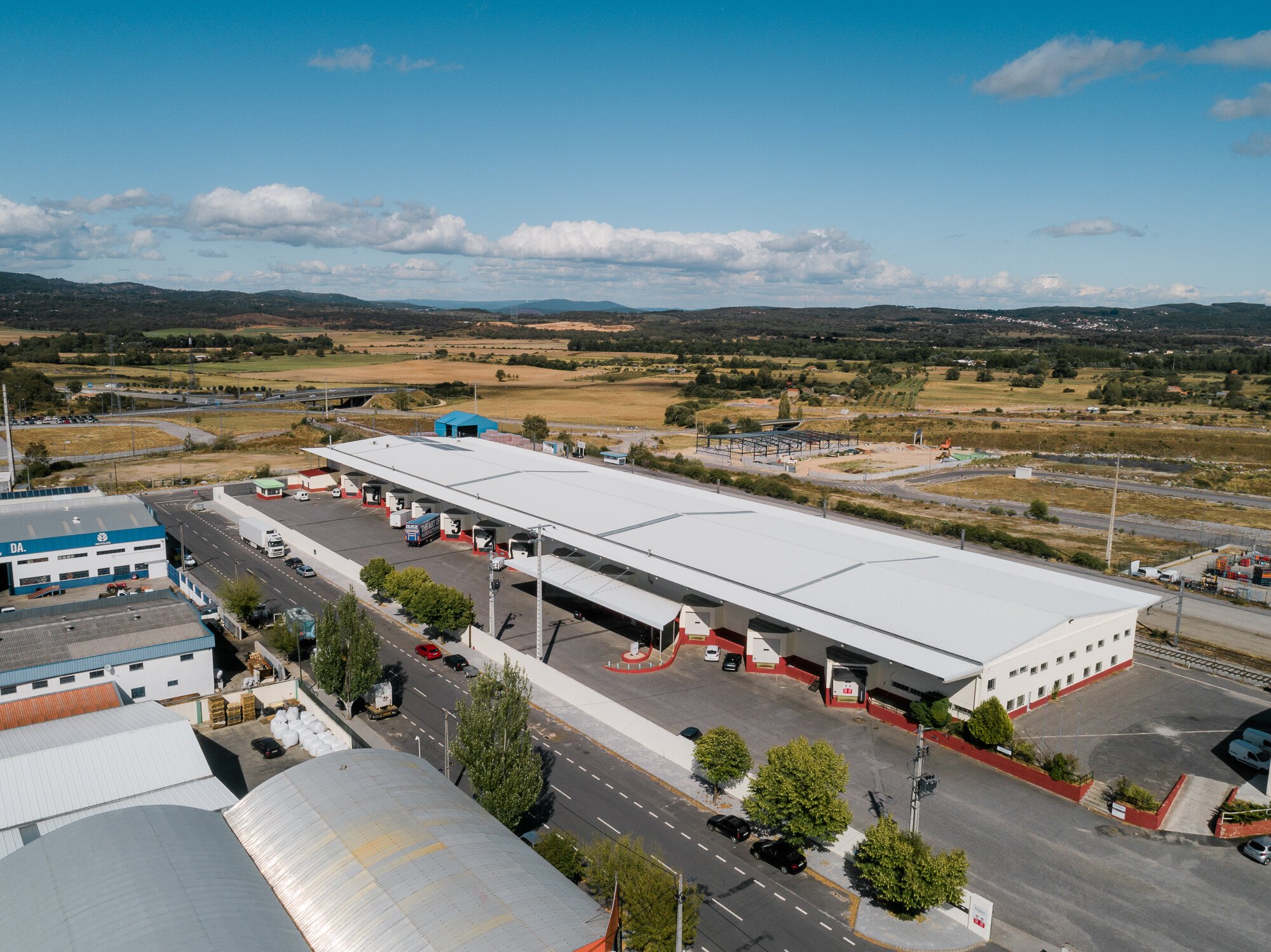 Vista aérea de um posto de combustível grande, com vários carros e caminhões, cercado por terrenos abertos e paisagens rurais ao fundo. Estradas pavimentadas e edifícios industriais estão próximos ao posto.