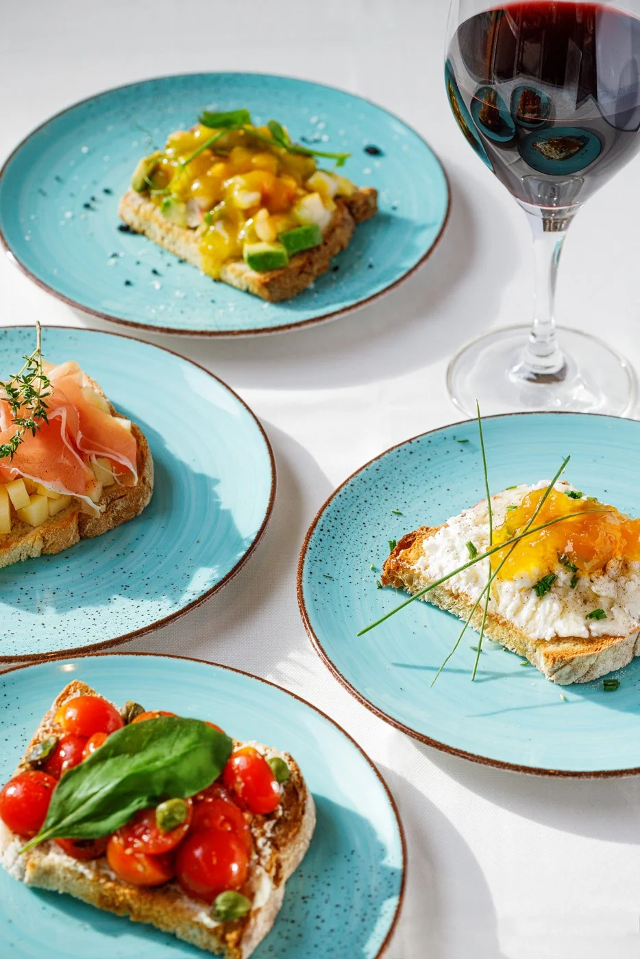 Toast decorado com ingredientes variados em pratos azuis e uma taça de vinho tinto ao lado.