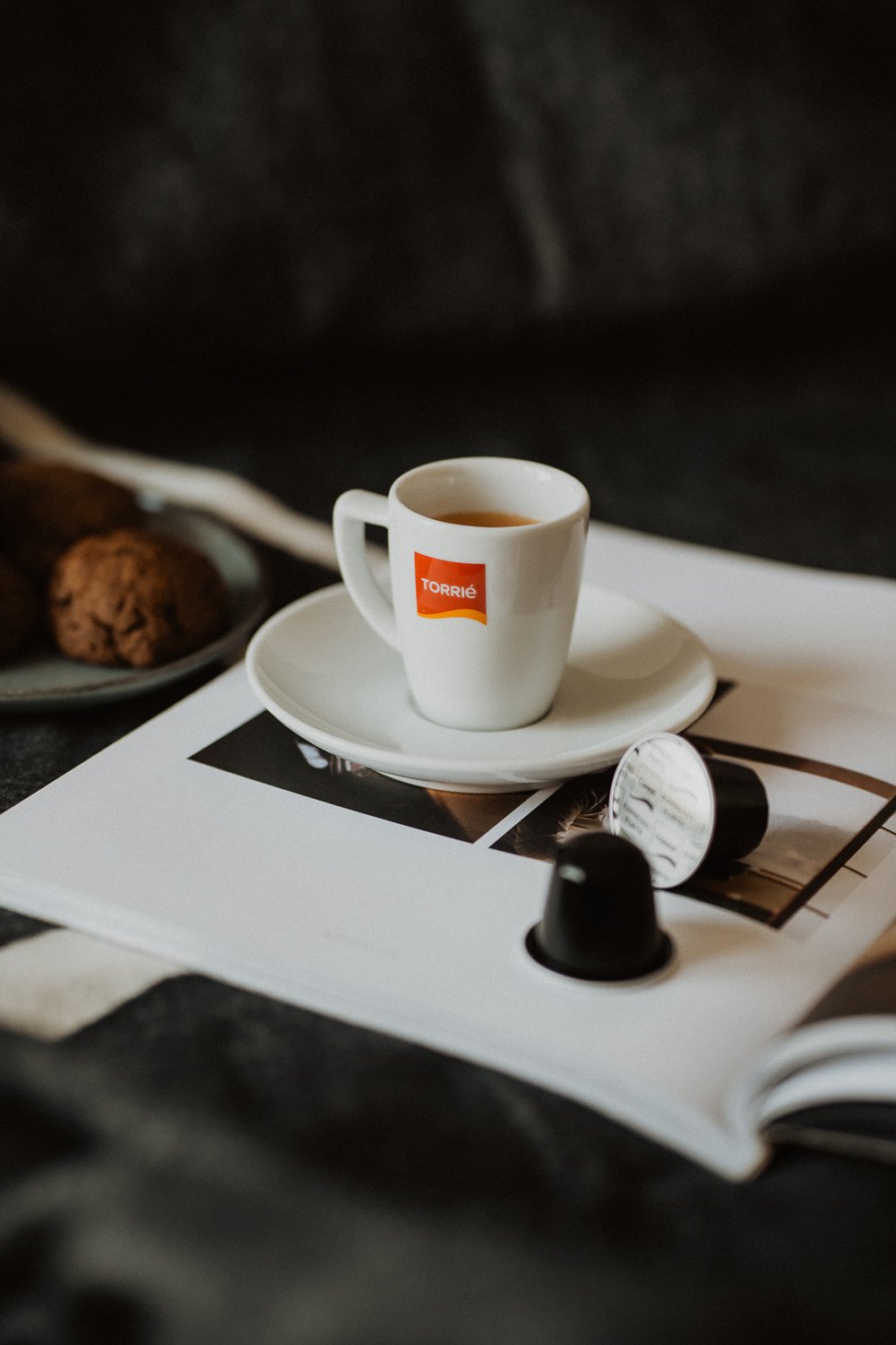 Xícara de café Torrie sobre pires com cápsulas de café ao lado e um prato de biscoitos ao fundo, tudo sobre uma revista aberta.