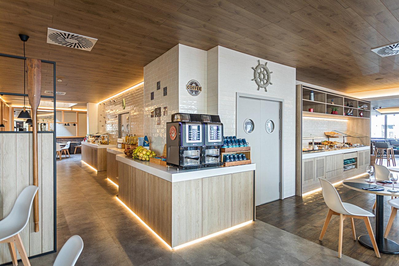 Área de café da manhã de um restaurante moderno, com balcão de café, frutas e pratos em bufê.