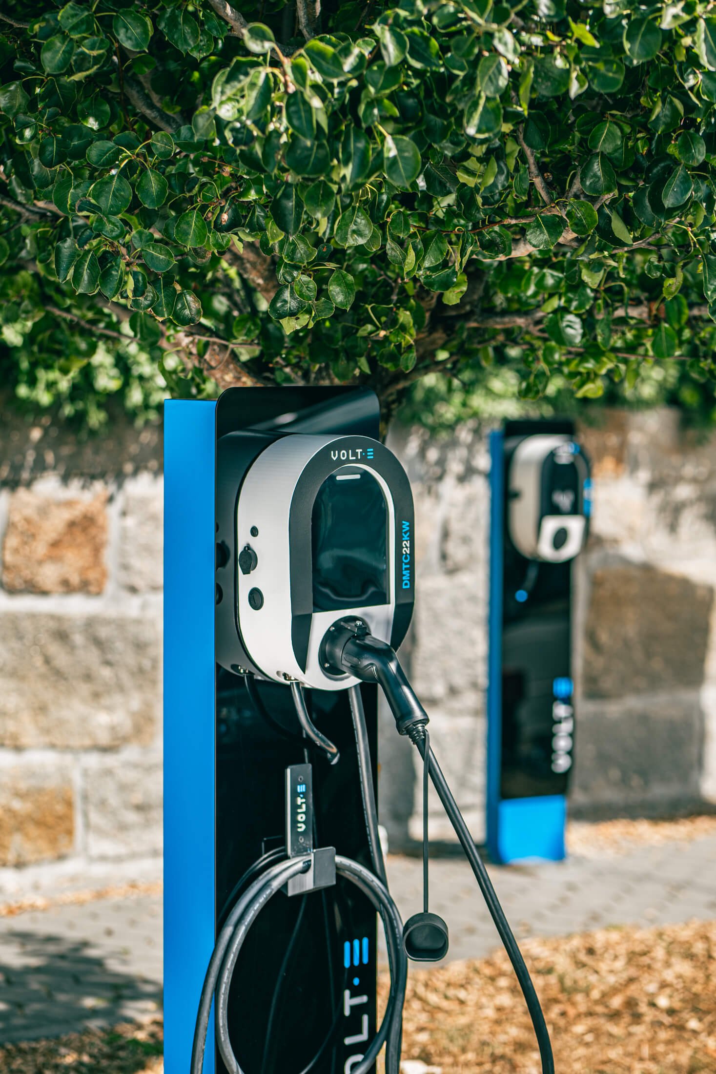 Estação de carregamento de veículos elétricos da marca Volt-e, com cabos conectados, localizada ao ar livre, cercada por vegetação e uma parede de pedra ao fundo.
