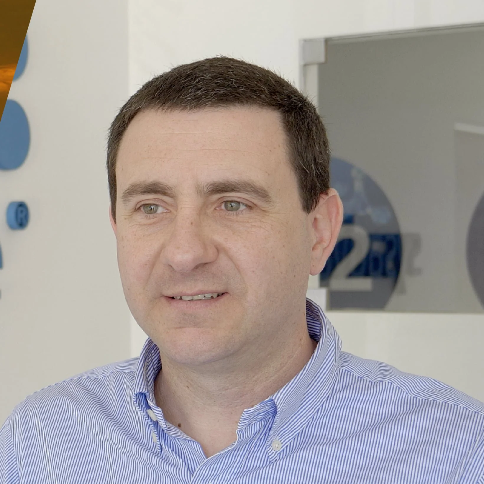Homem em camisa listrada azul, em ambiente de escritório com parede branca e decoração azul. Texto parcial "2" visível no fundo.