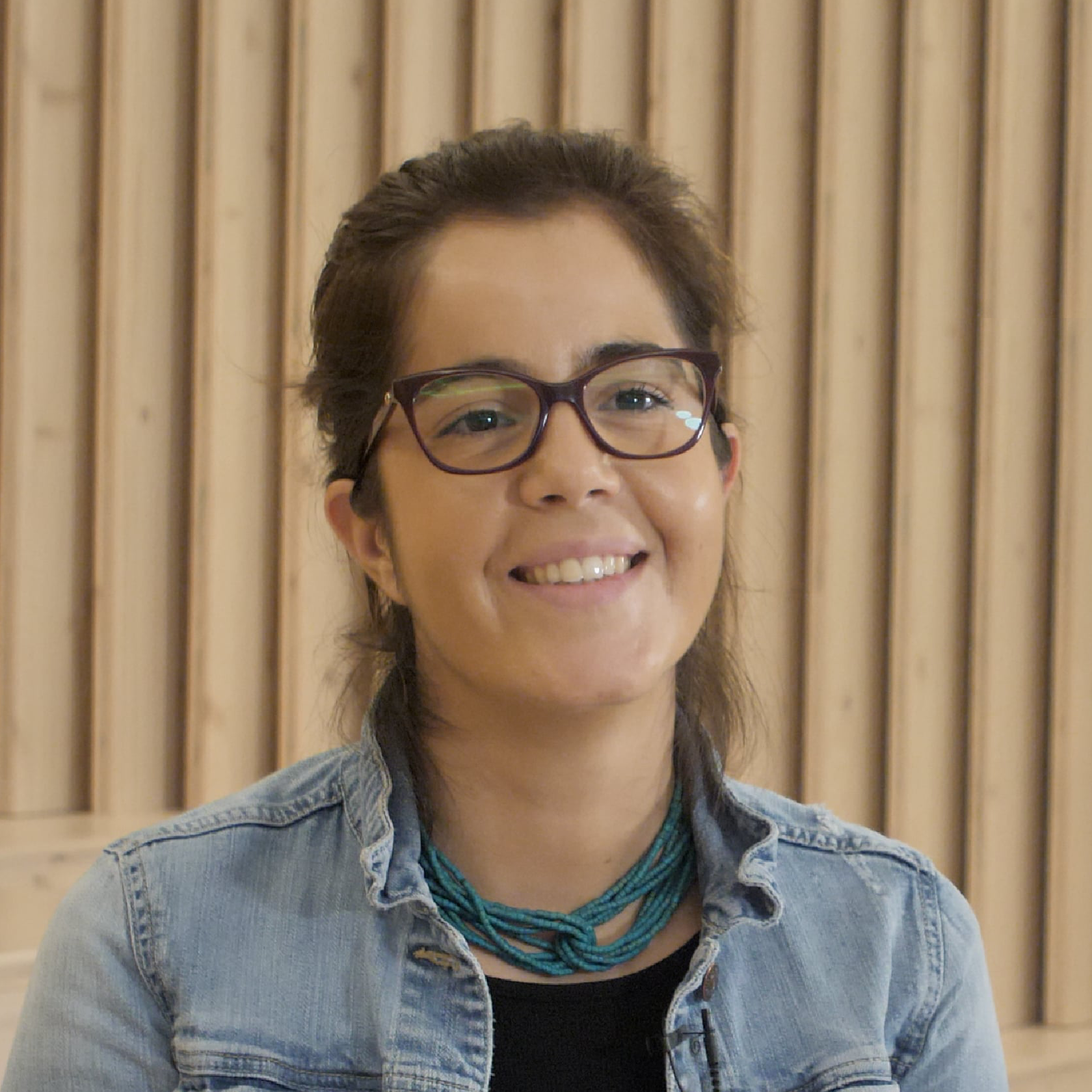 Mulher de óculos sorrindo, vestindo jaqueta jeans e colar azul, com fundo de madeira.