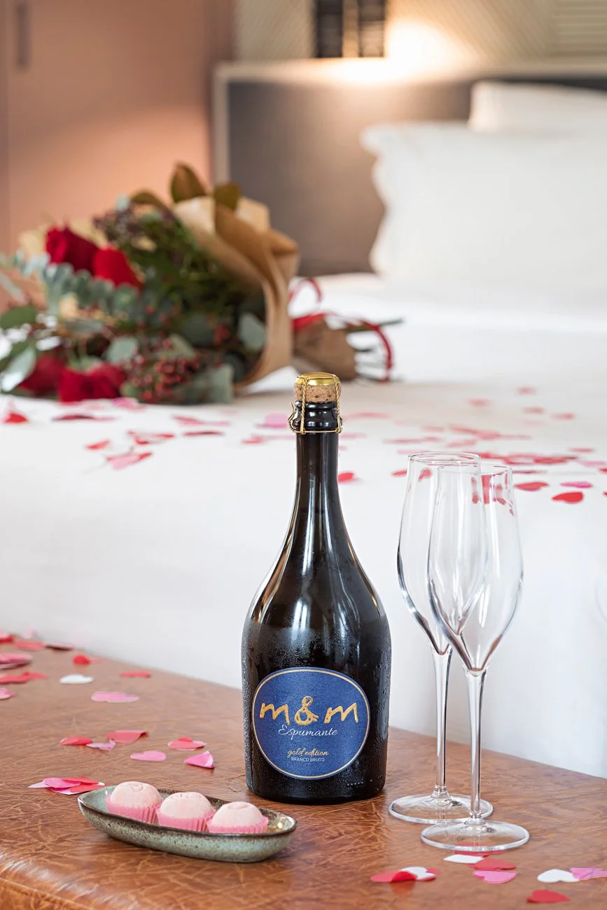 Garrafa de espumante M&M sobre uma mesa, acompanhada de duas taças vazias. Ao fundo, pétalas de rosa espalhadas em uma cama e um buquê de flores.