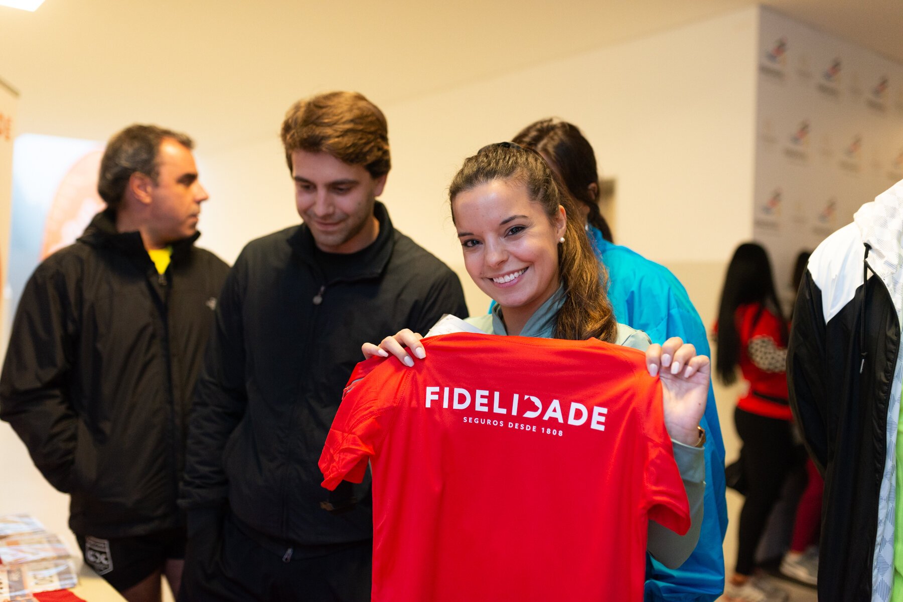 Mulher sorridente segurando camiseta vermelha com a palavra "Fidelidade". Outras pessoas ao fundo em um ambiente interno.