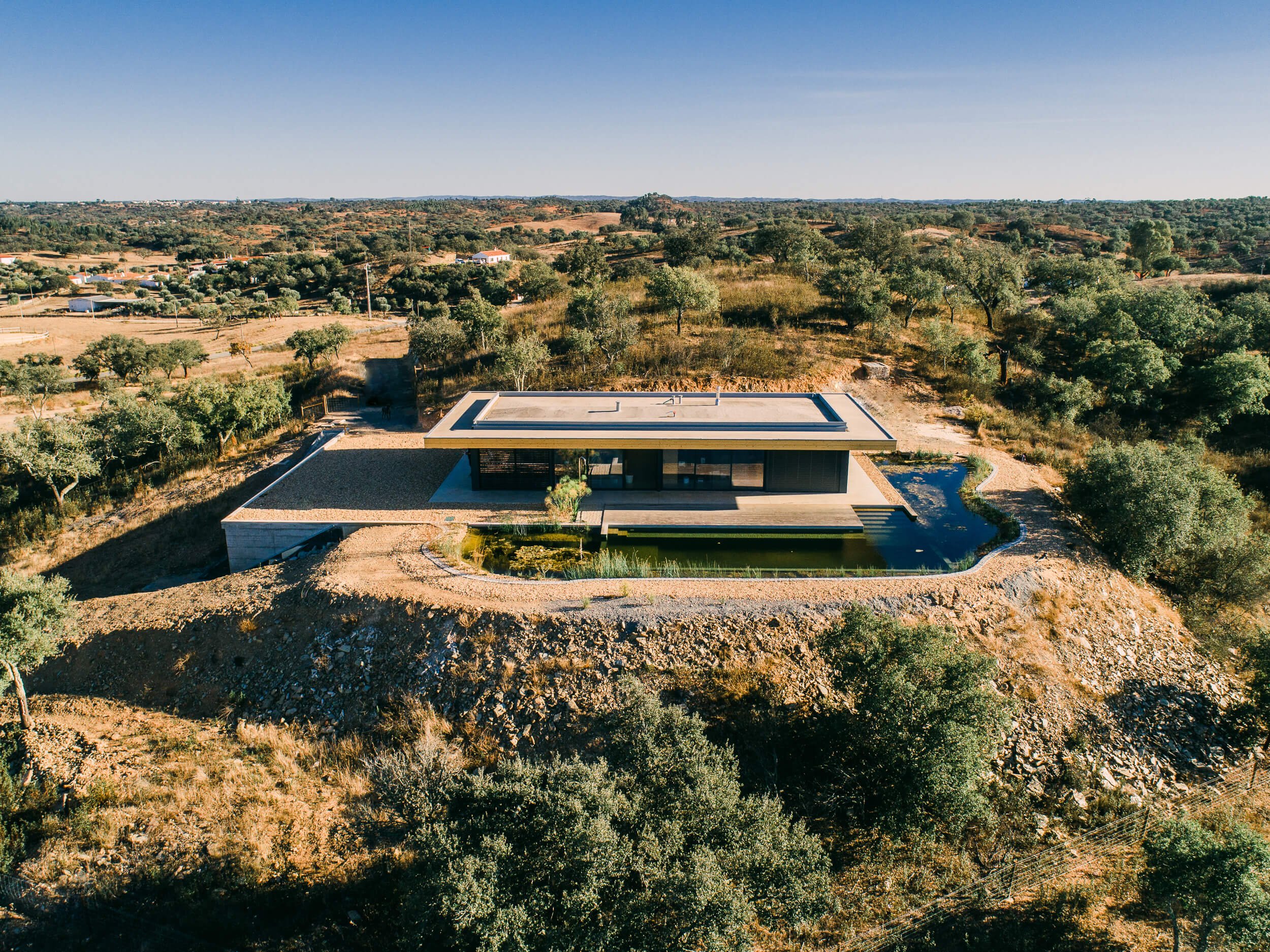 Casa moderna em área rural, cercada por árvores e terreno seco, com piscina ao redor, sob céu azul sem nuvens.